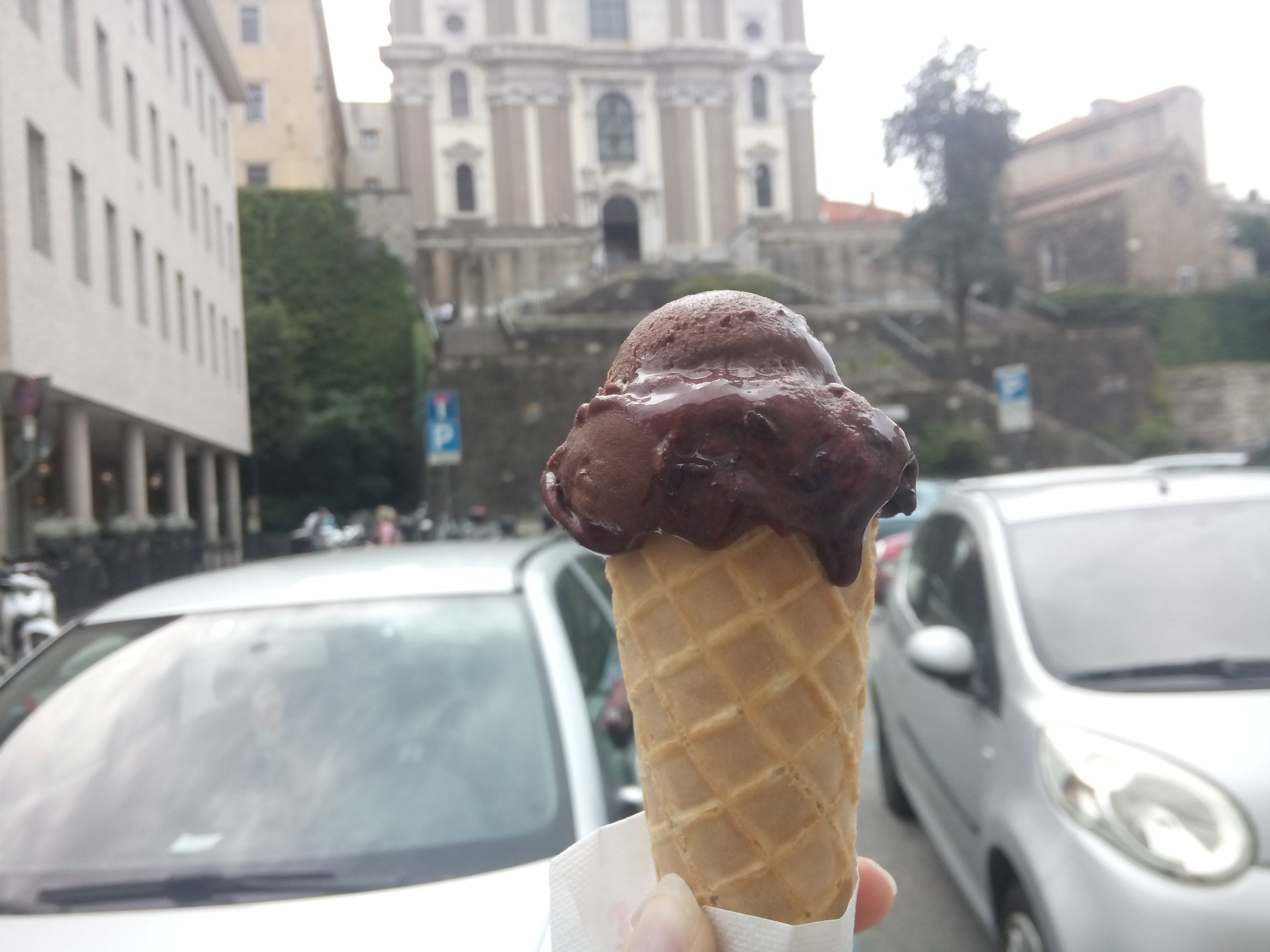 Chocolate ice cream in a cone in the foreground, with cars and a historical building in the background