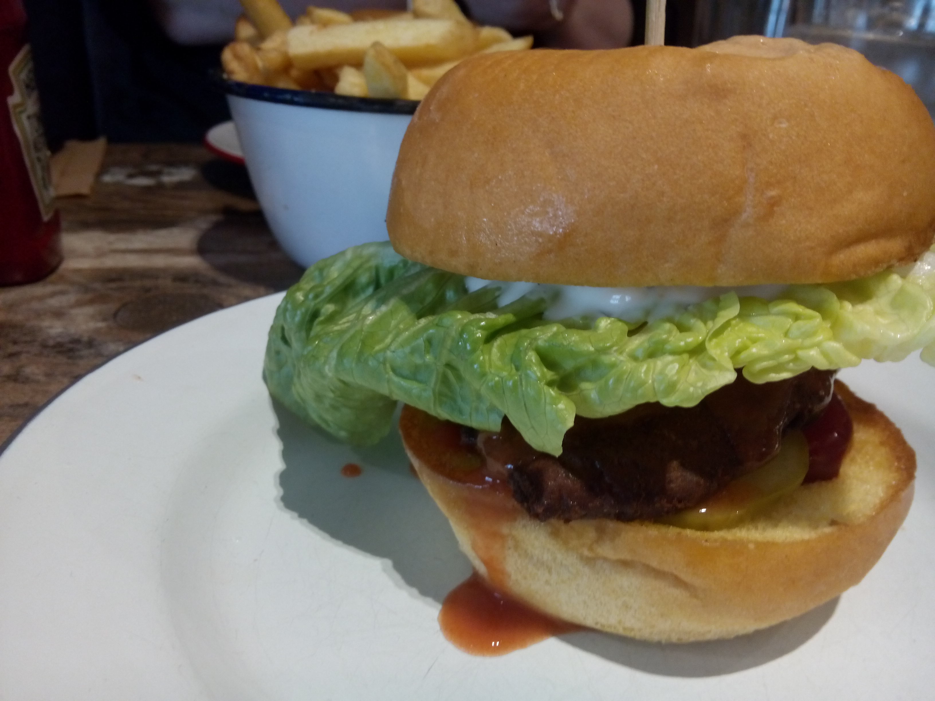A seitan burger in a bun on a white plate, with salad and blue cheese sauce poking out; chips in the background