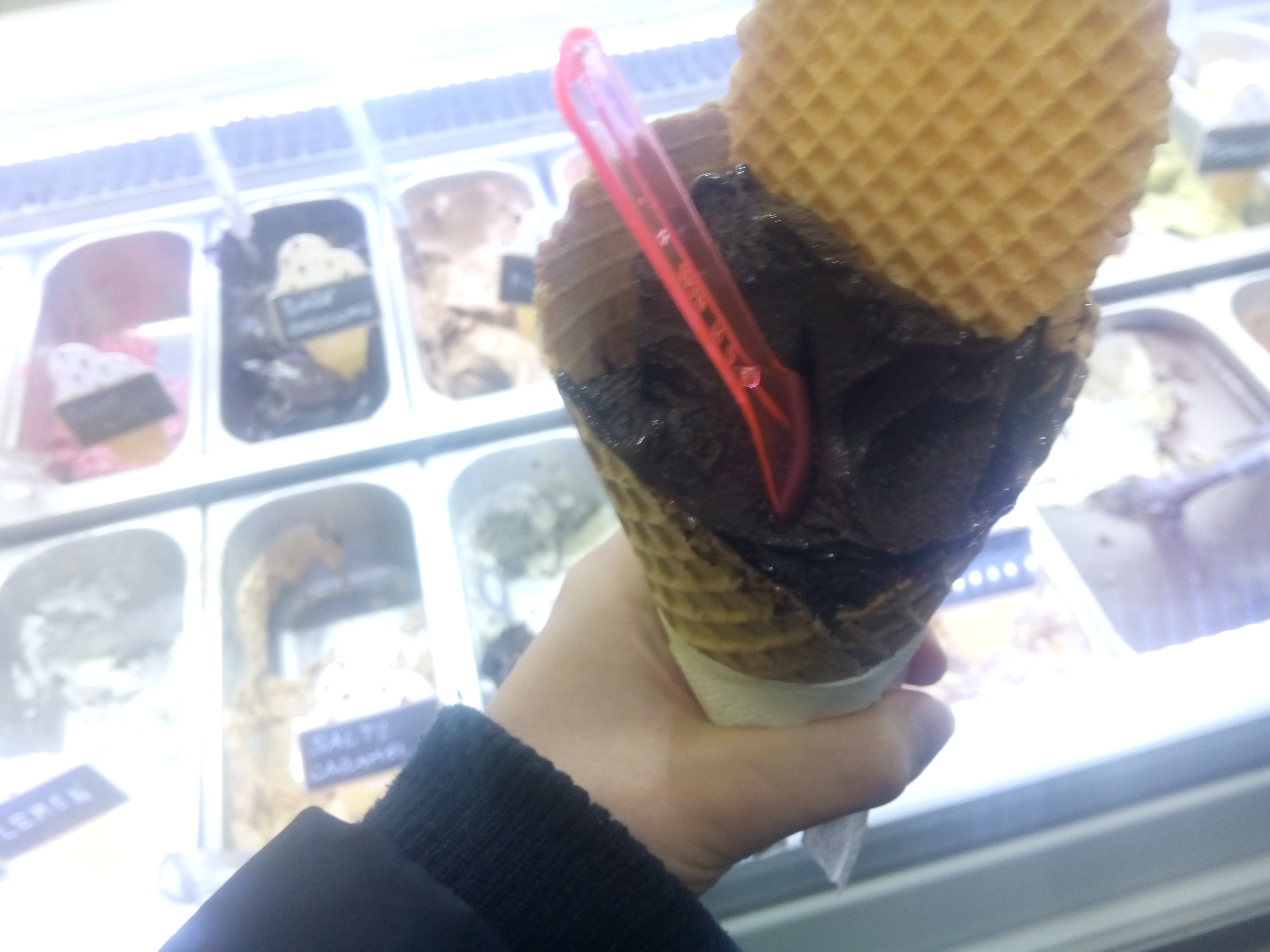 A large dark chocolate ice cream in a waffle cone, against a background of more ice cream