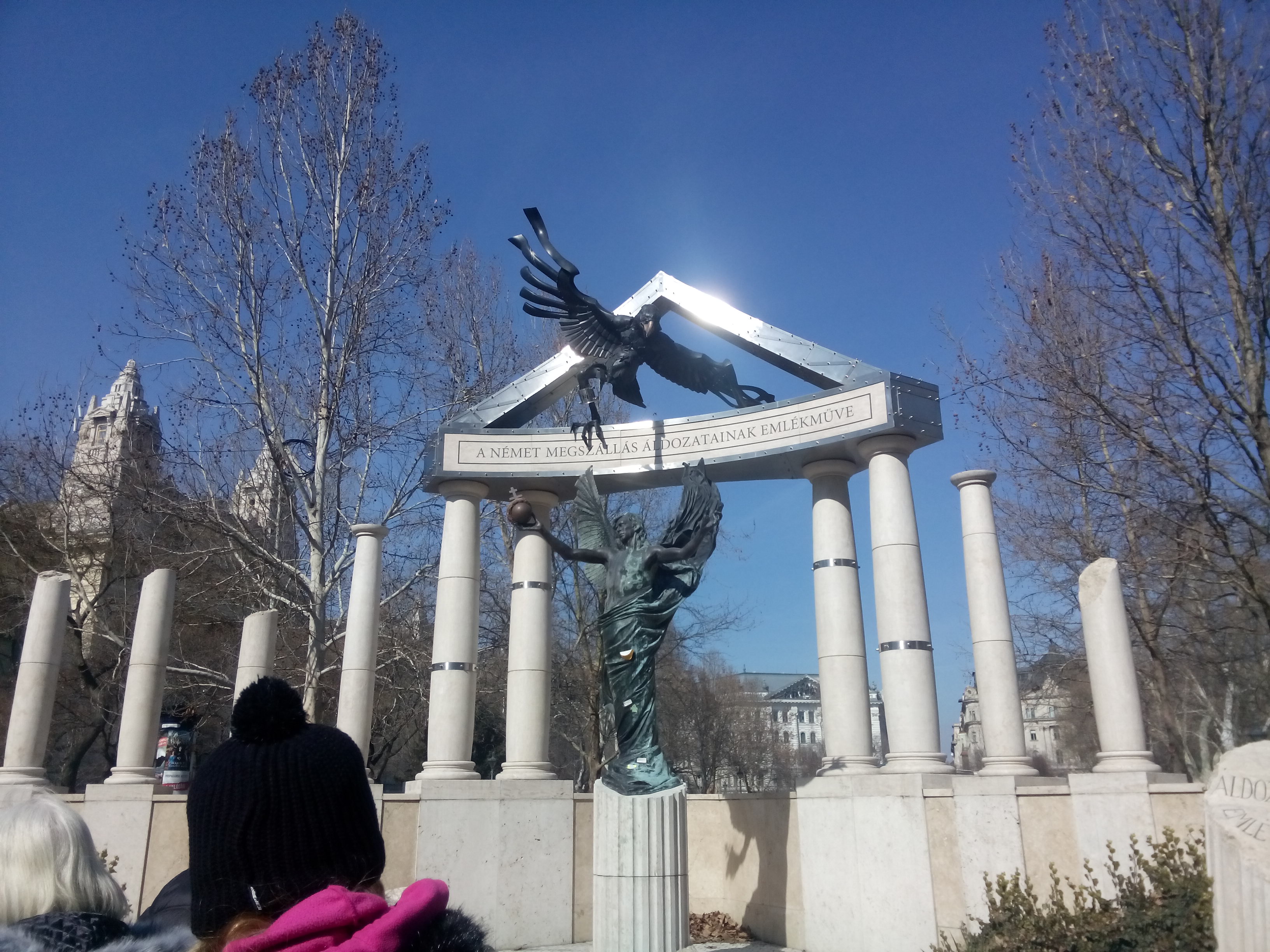 A metal statue of an eagle on top of white stone pillars against ablue sky with tress