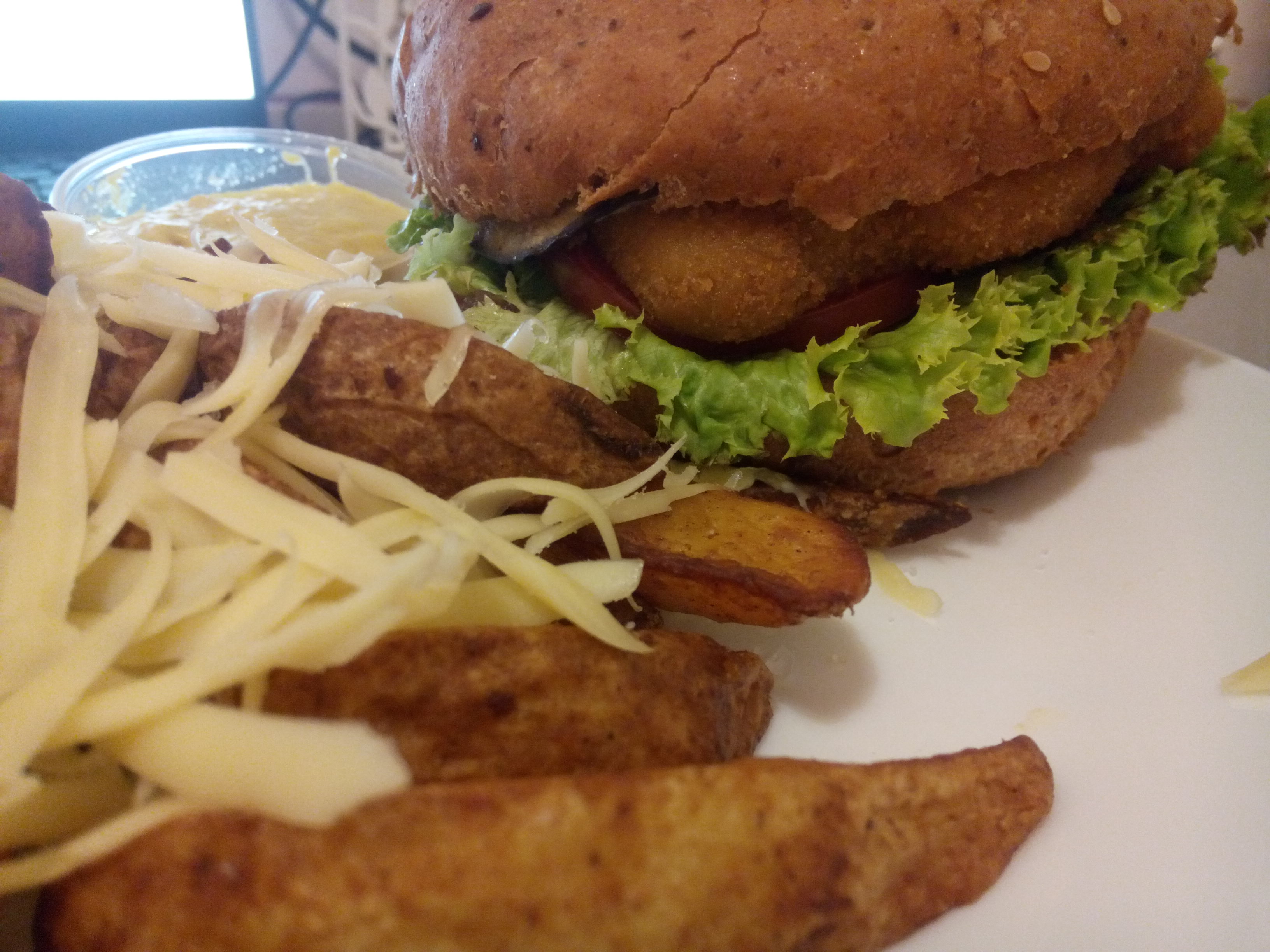 A close up on a pile of potato wedges covered with grated cheese, beside a burger pun out of which pokes frilly lettuce, tomato, and a breaded patty