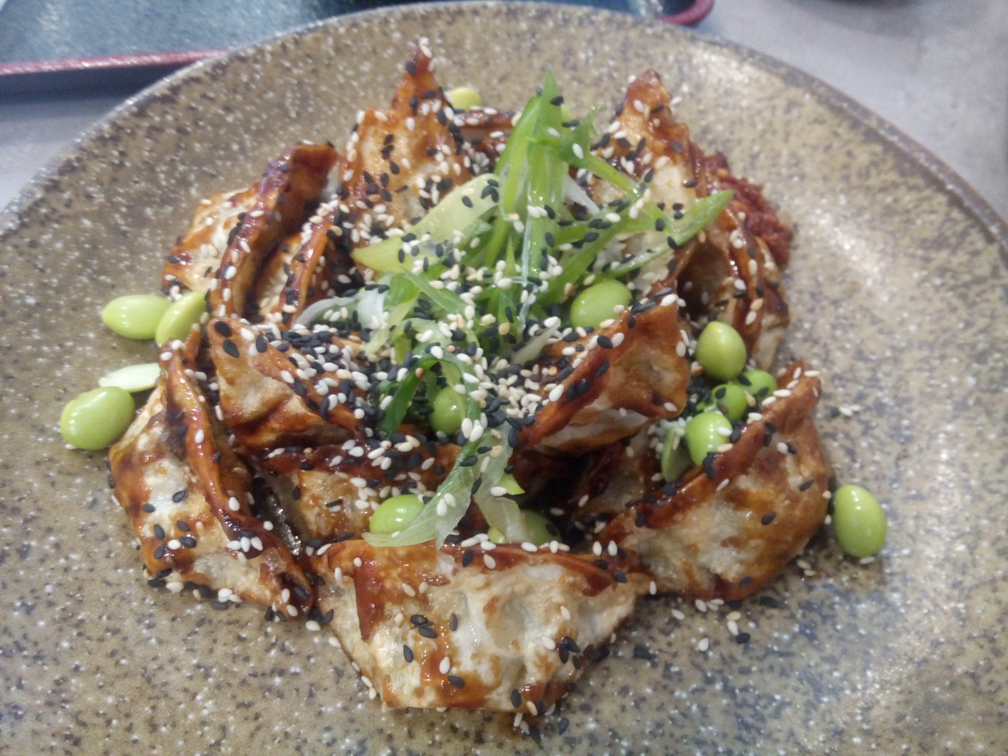 A speckled brown dish with a pile of dumplings, scattered with green soya beans, garnish and sesame seeds