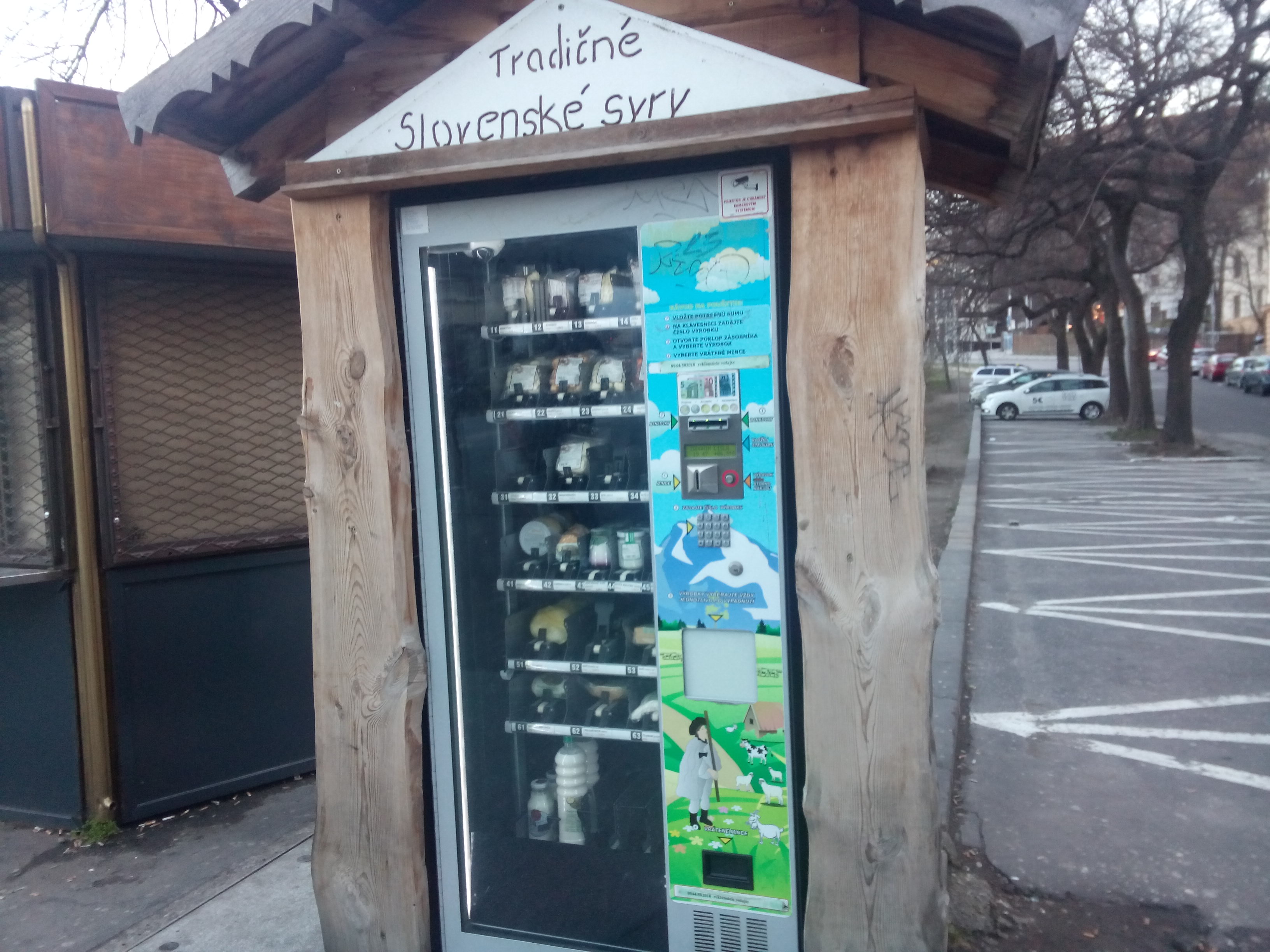 A wooden frame around a modern vending machine, containing traditional Slovakian cheeses