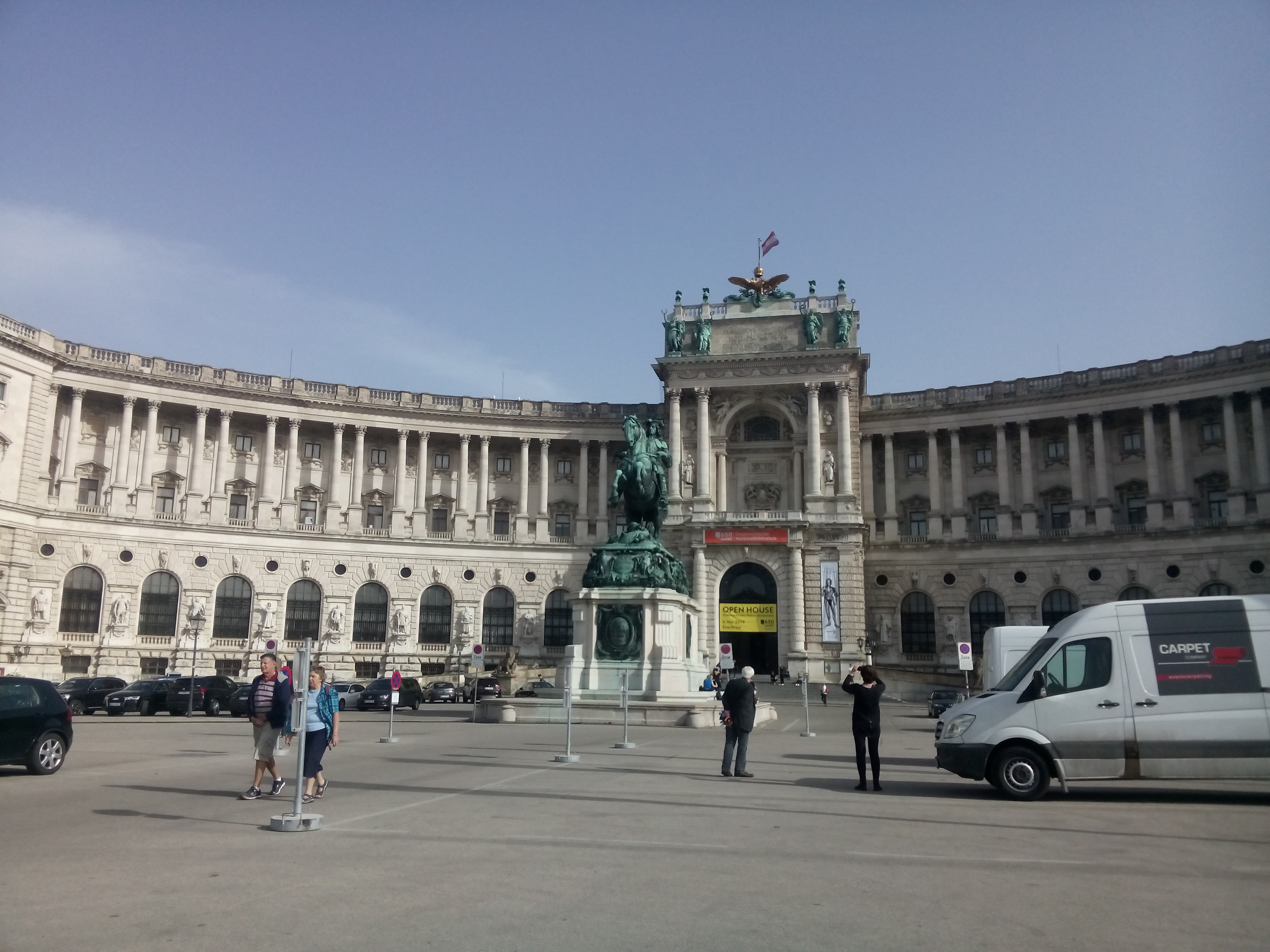 Curved grand building with a green statue, tarmac in front, some cars and tourists