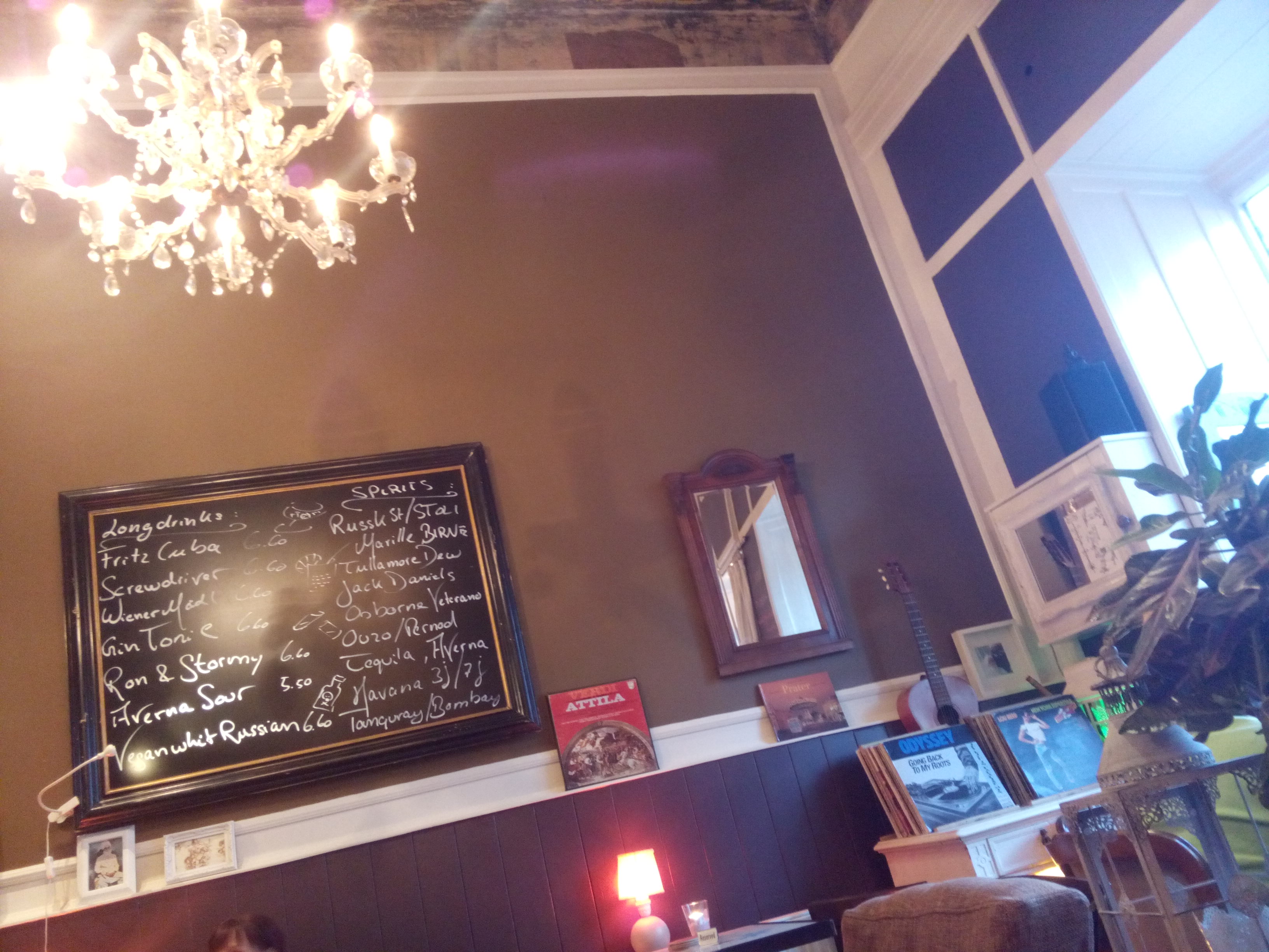 Cafe interior, with a chandelier in the top left, a blackboard with writing, brown walls and the tops of chairs