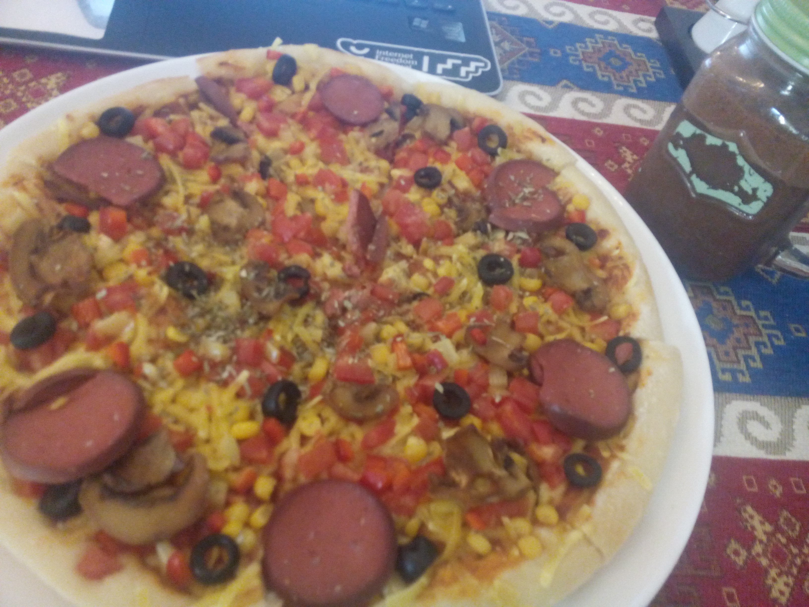 A colourful pizza on a plate with various veggies and not-sausage, on a table with a chocolate-coloured glass