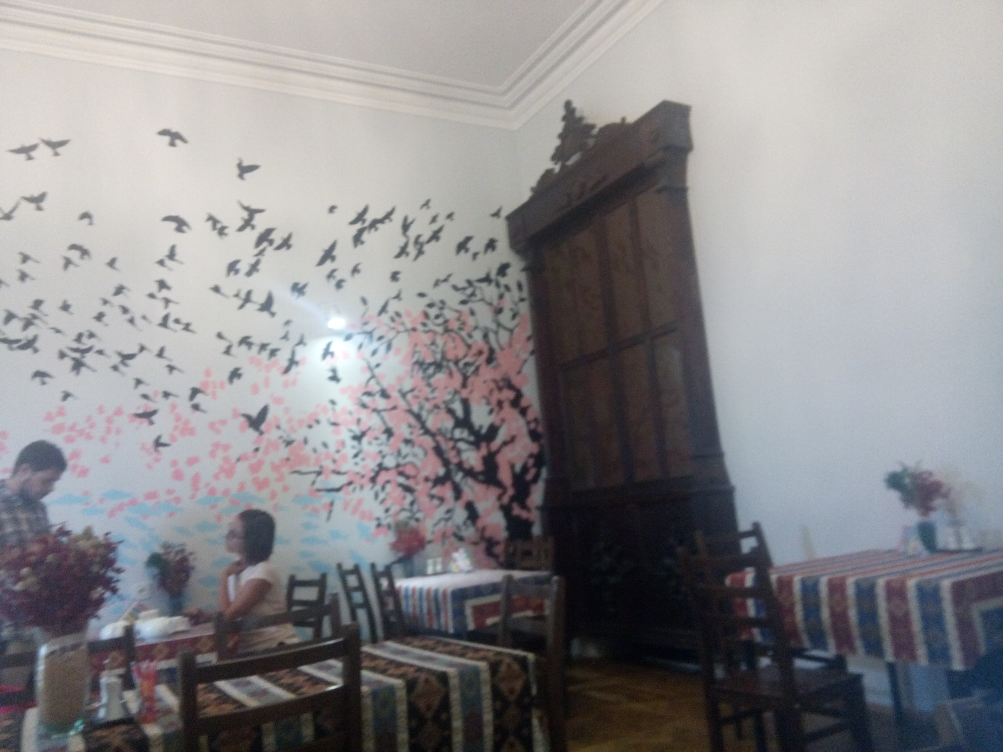 Inside a cafe with several tables, an ornate wooden cabinate against the wall, and a mural of birds flying from a tree