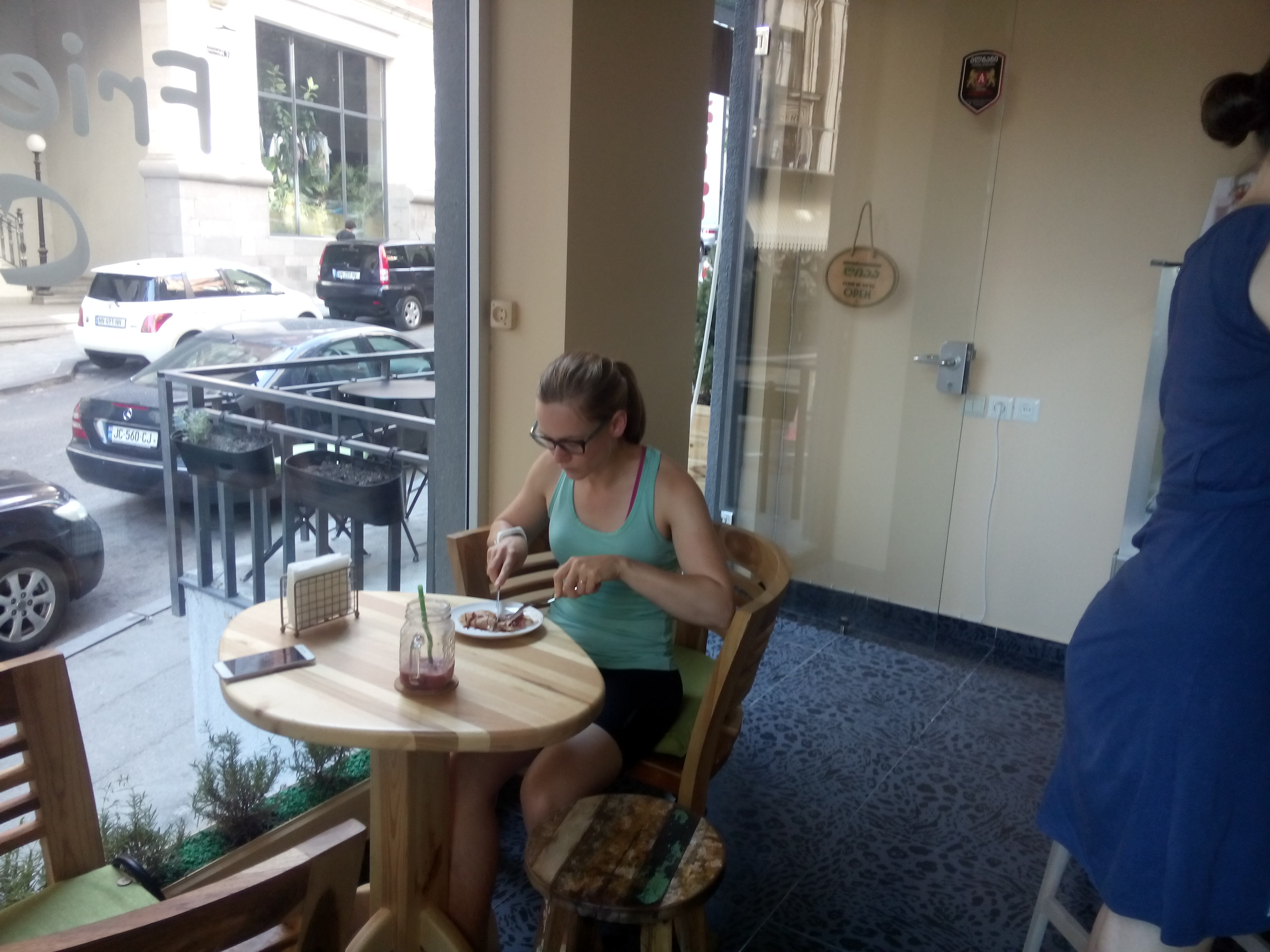 A woman sits at a table by a window in a cafe, eating a pancake with a knife and fork