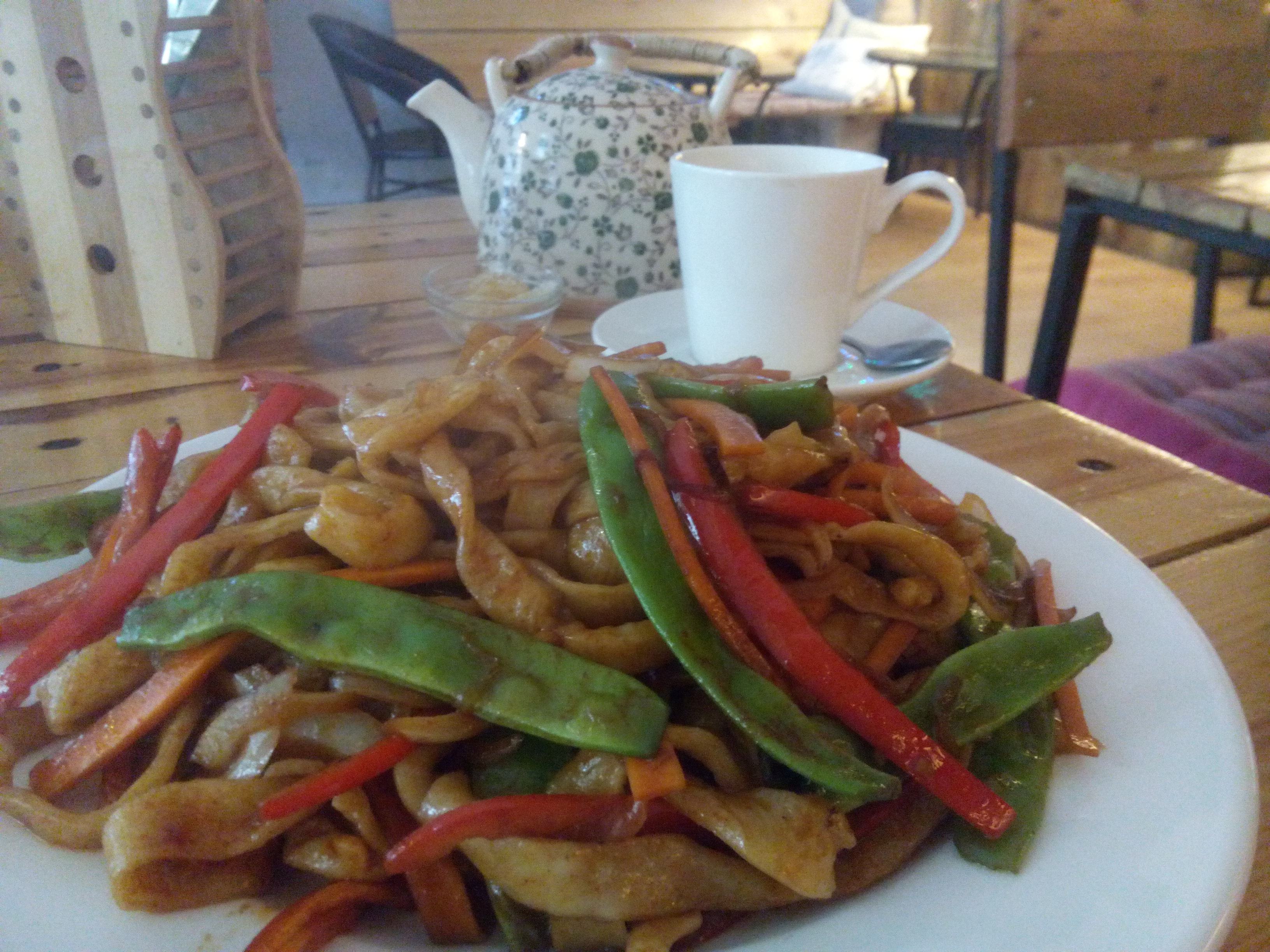A plate piled high with brown noodles, red and green vegetables, and a teacup and pot in the background