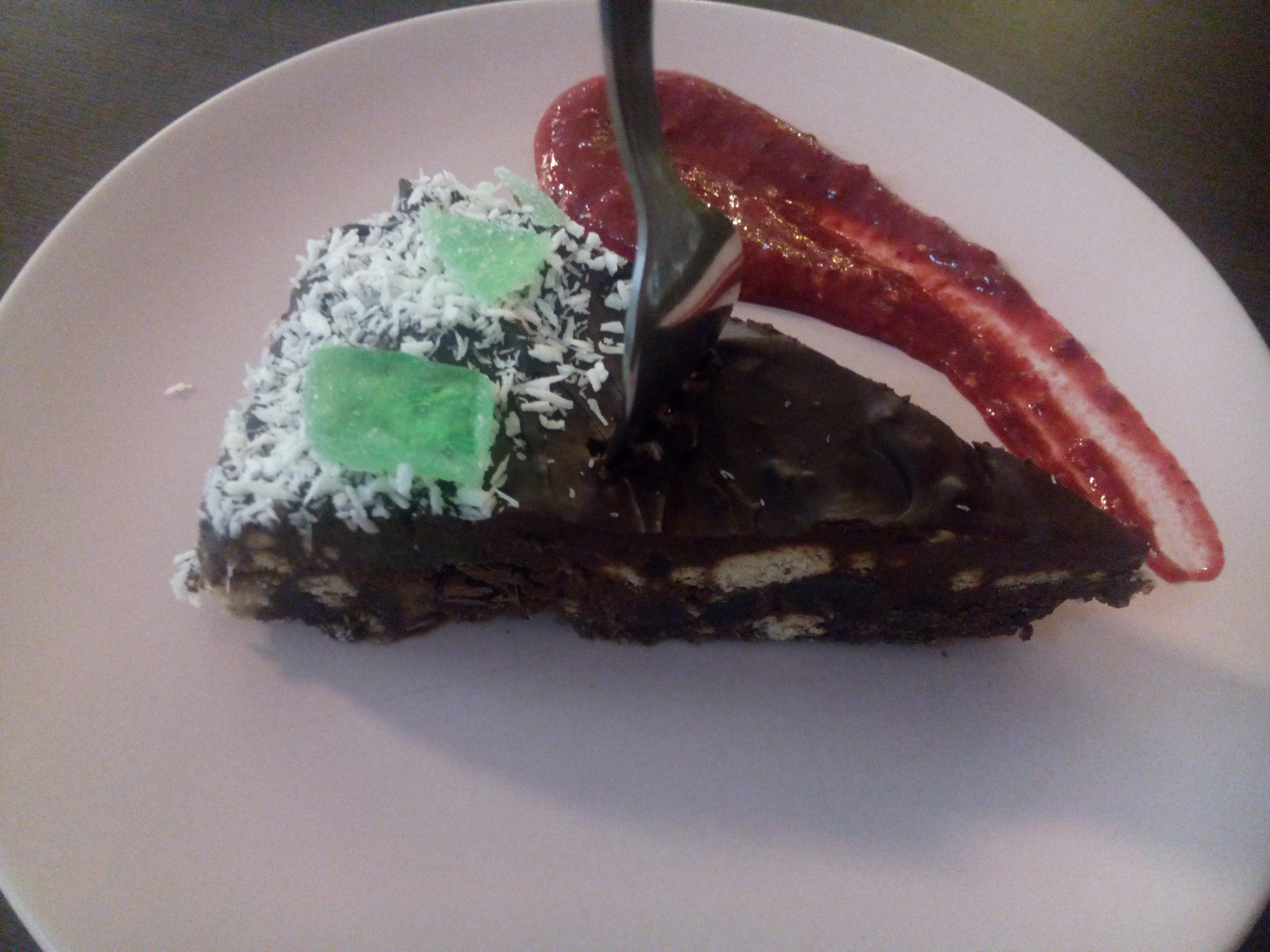 A white cake with a smear of red jam around a triangle of cake, with coconut sprinkles and green candy on top, and a fork stuck upwards
