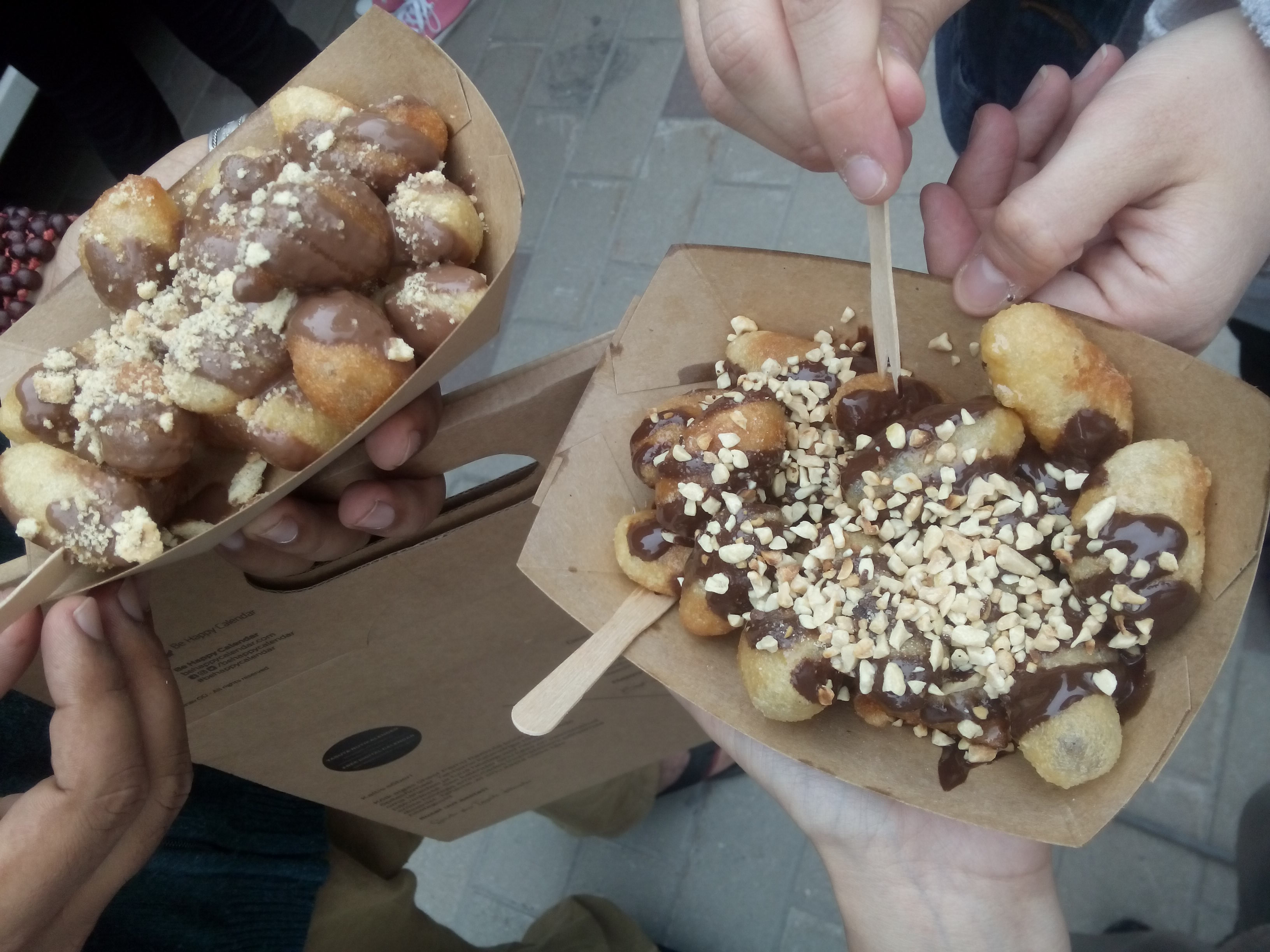 Hands holding two cardboard trays of donut holes covered in chocolate and nuts