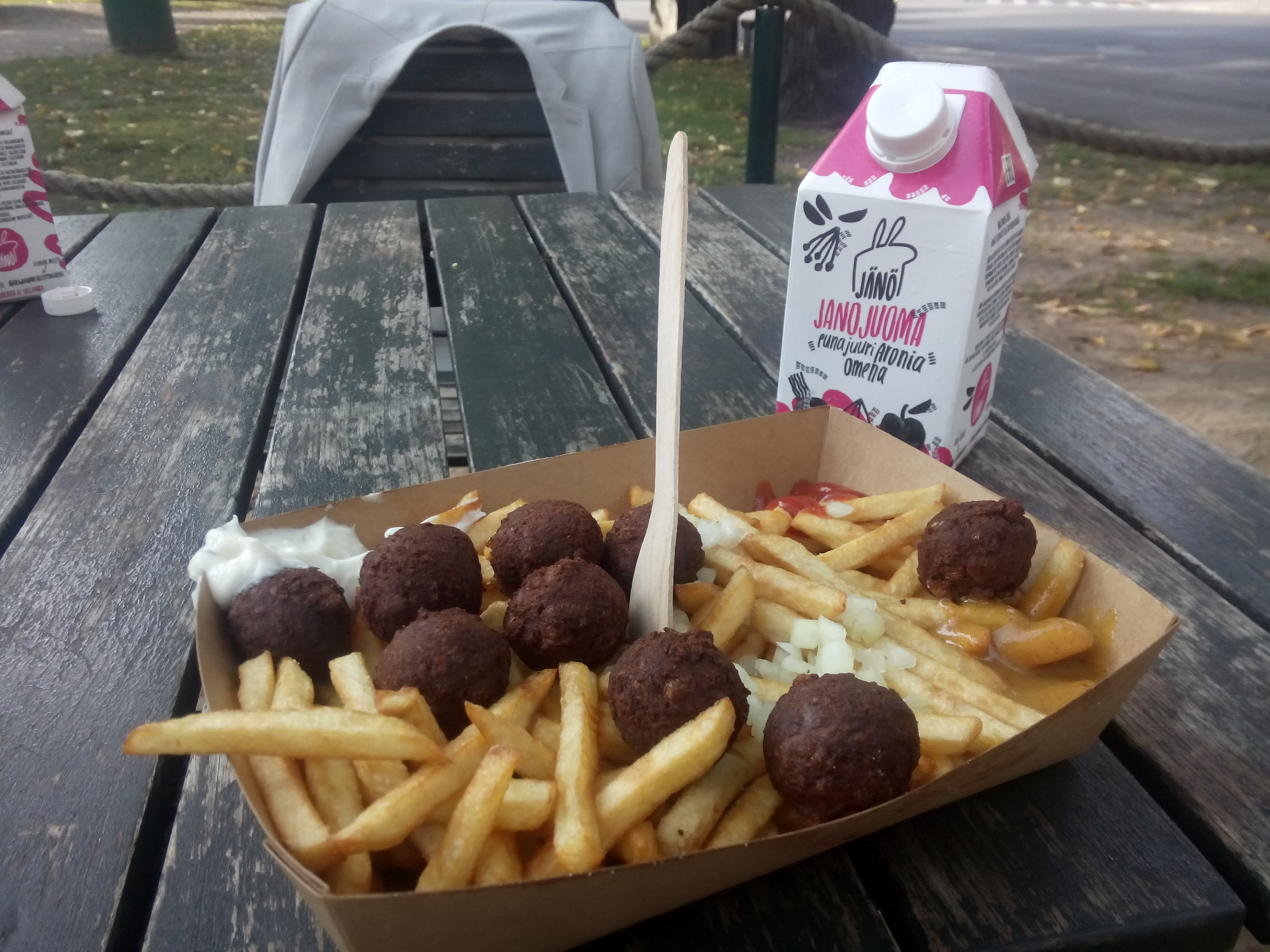 A wooden table outside holds a cardboard tray containing thin fries and not-meatballs; a pink and white carton of juice beside it