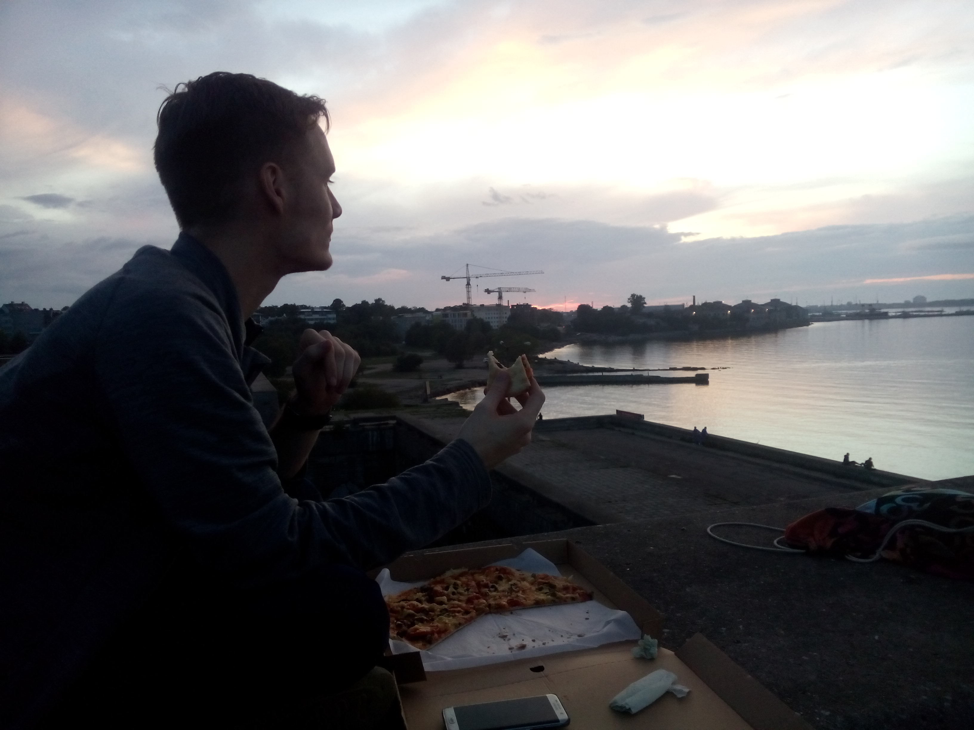 A guy with short hair eats pizza from a box looking out of water and sunset