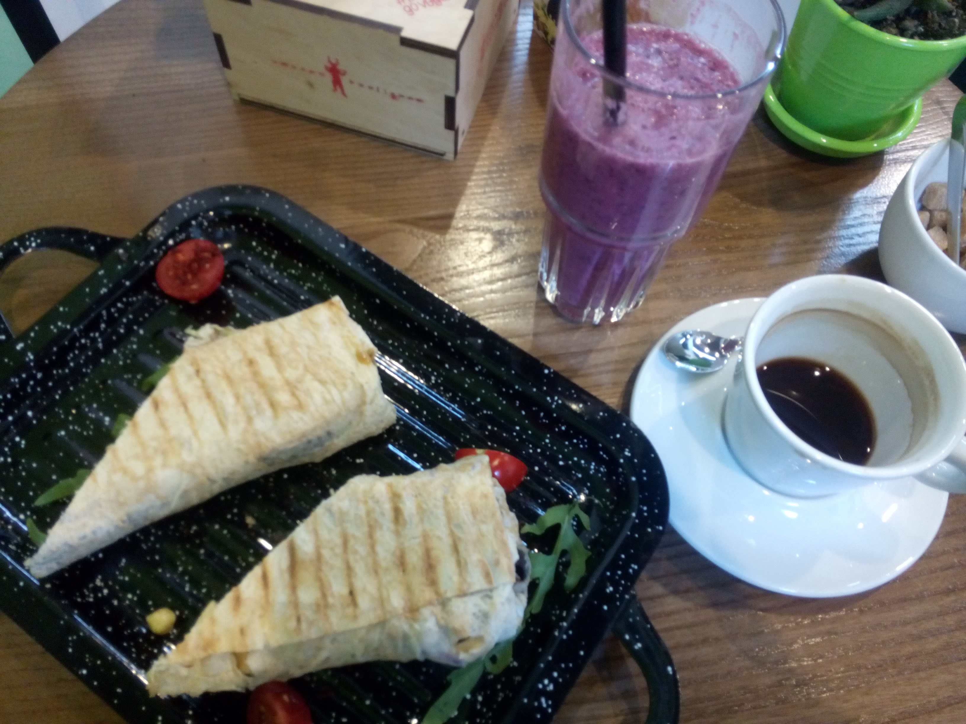 Two halves of a tortilla wrap on a black tray, on a table beside a purple smoothie in a glass and an espresso in a white cup and saucer