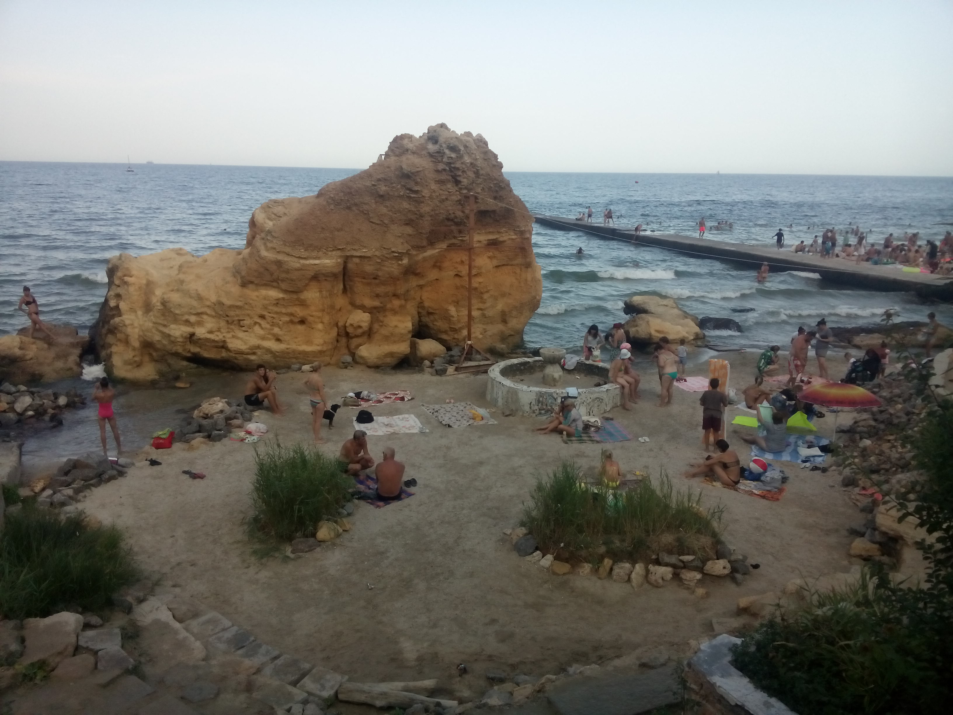 An area of beach with people sitting around and a large rock on the edge of the sea