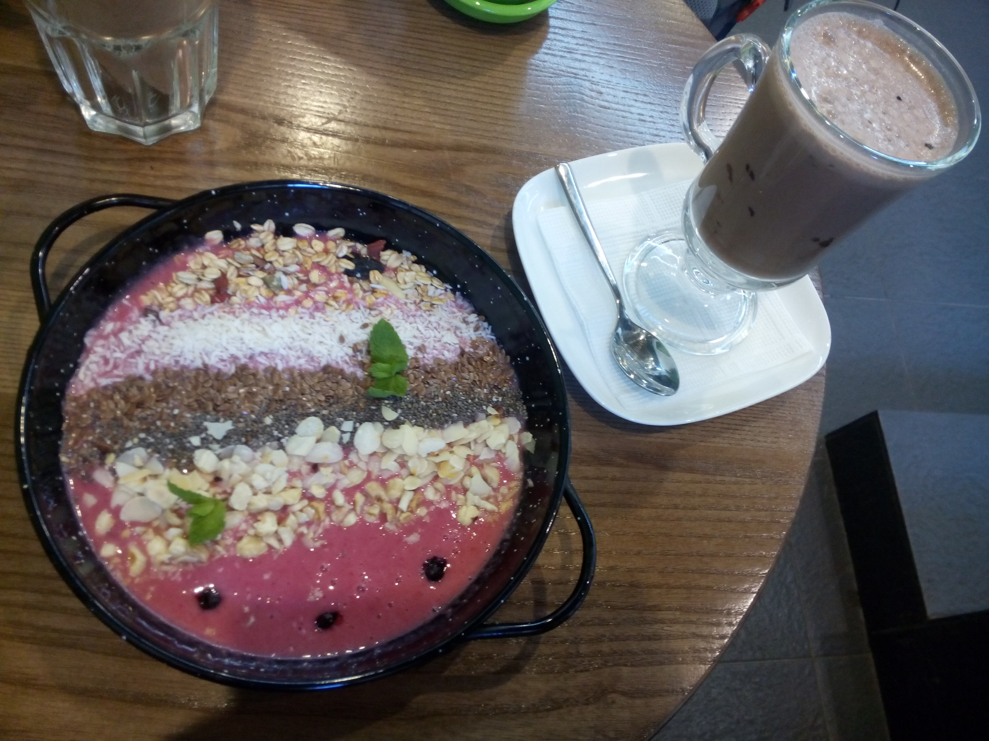 A wooden table with a black bowl containing smoothie, nuts and seeds, beside a hot chocolate in a tall glass