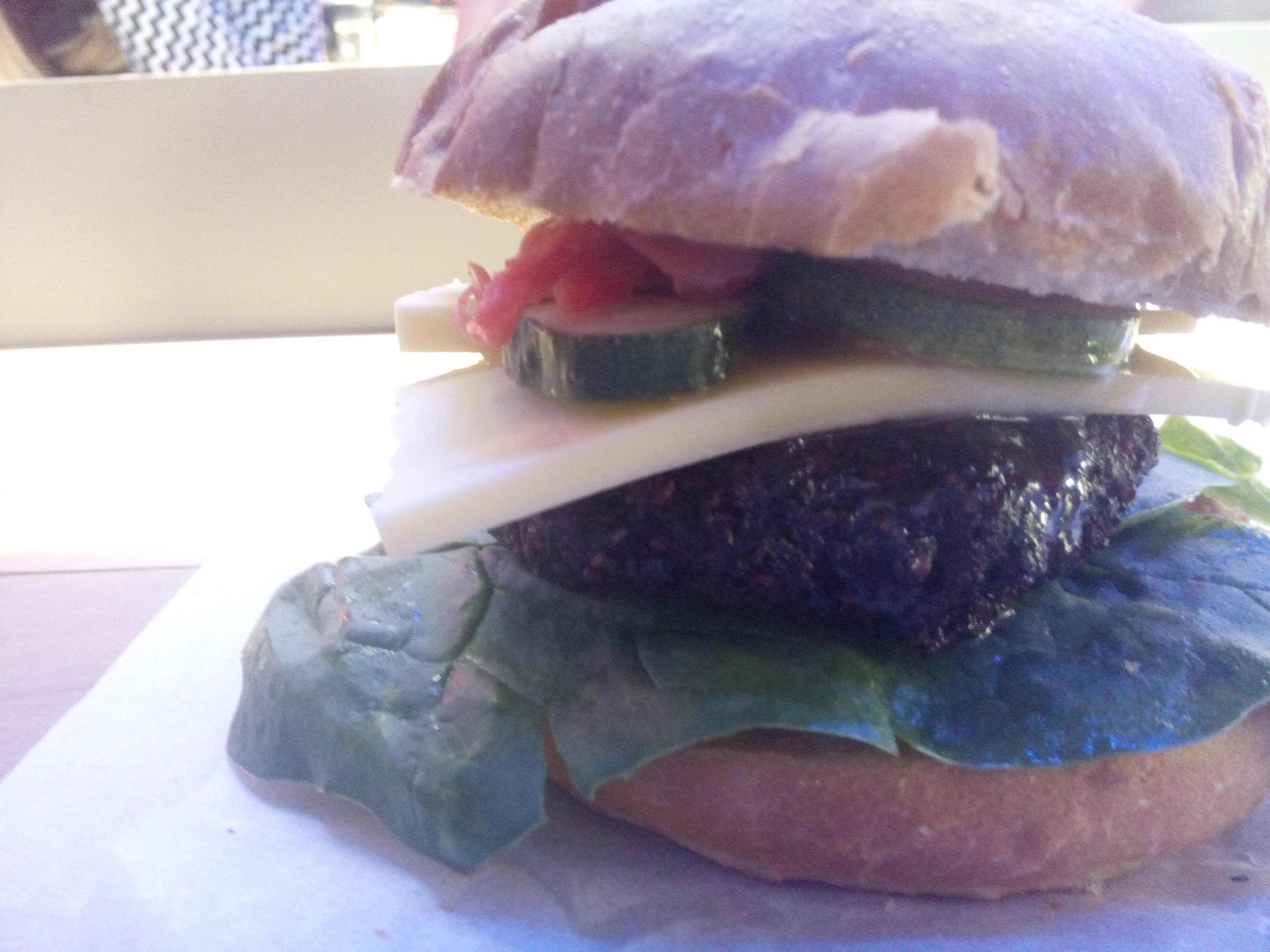 Close up side view of a burger tower; bread with spinach, patty, thick slice of vegan cheese, pickles and salad
