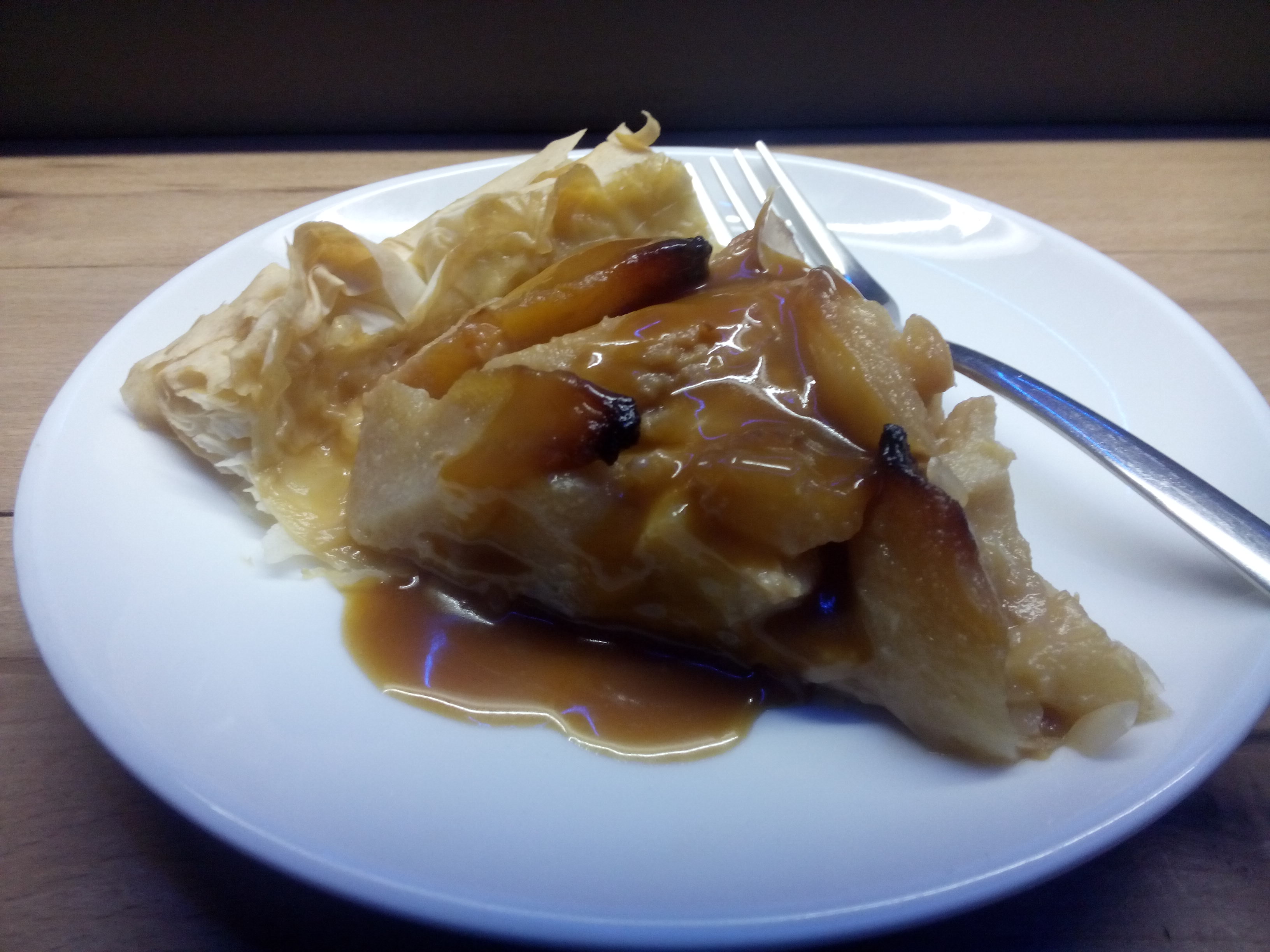 Flasky pastry tart on a white plate with a fork. The tart is full of pear and drizzled with shiny caramel sauce