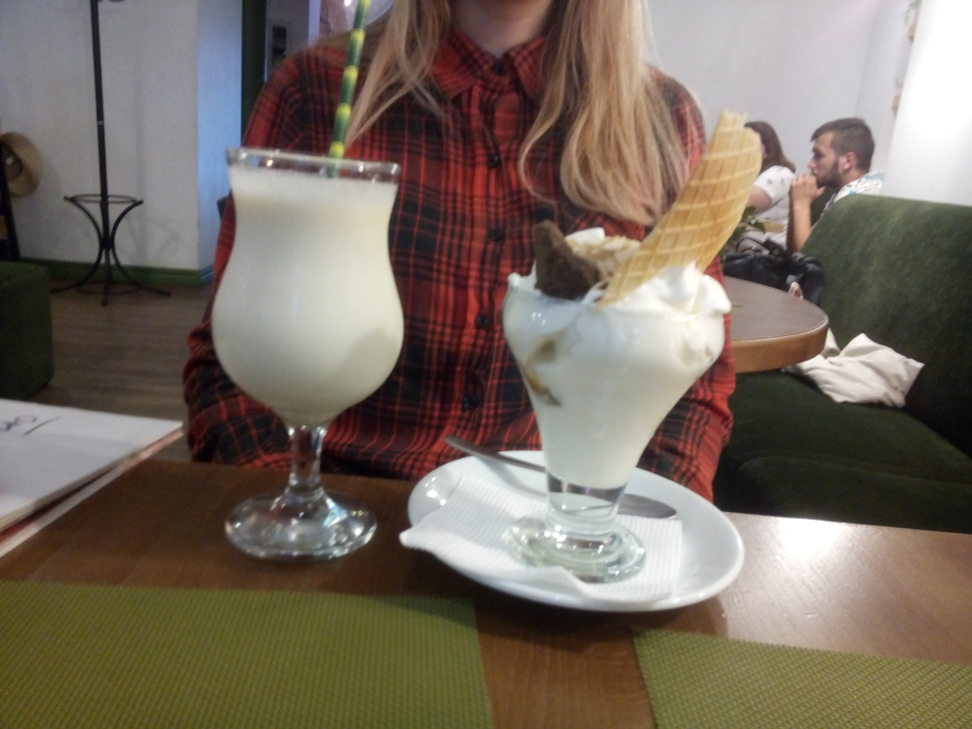 The torso of a woman in a red check shirt is visible behind a large cocktail glass full of something creamy and an icecream sundae with chocolate and wafers