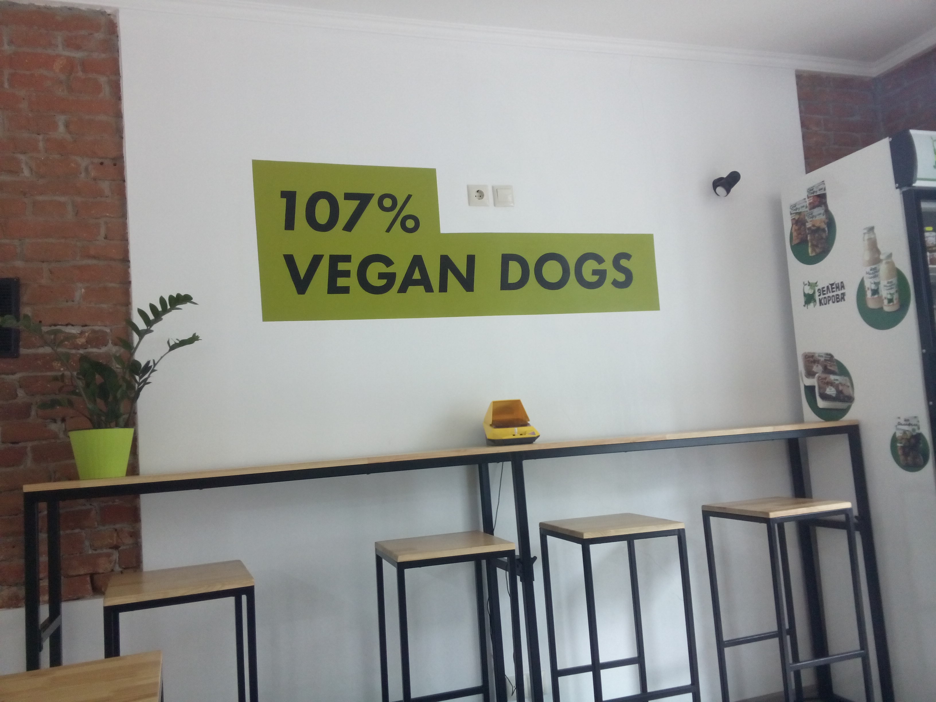 A white and brick wall with a shelf and high stools, and a green painted sign saying '107% vegan dogs'