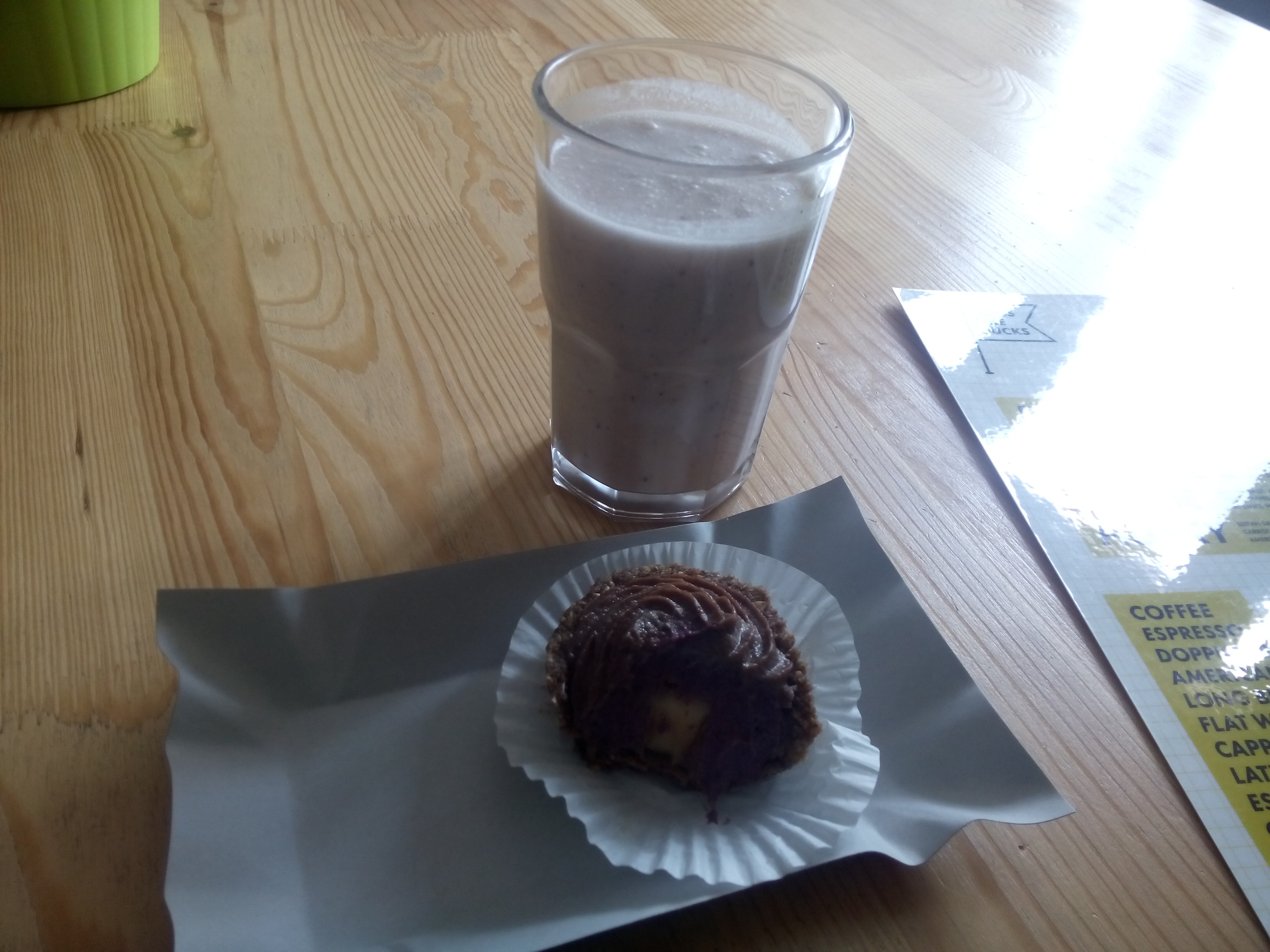 A wooden table with a glass of milkshake and a small chocolatey cake on cardboard tray