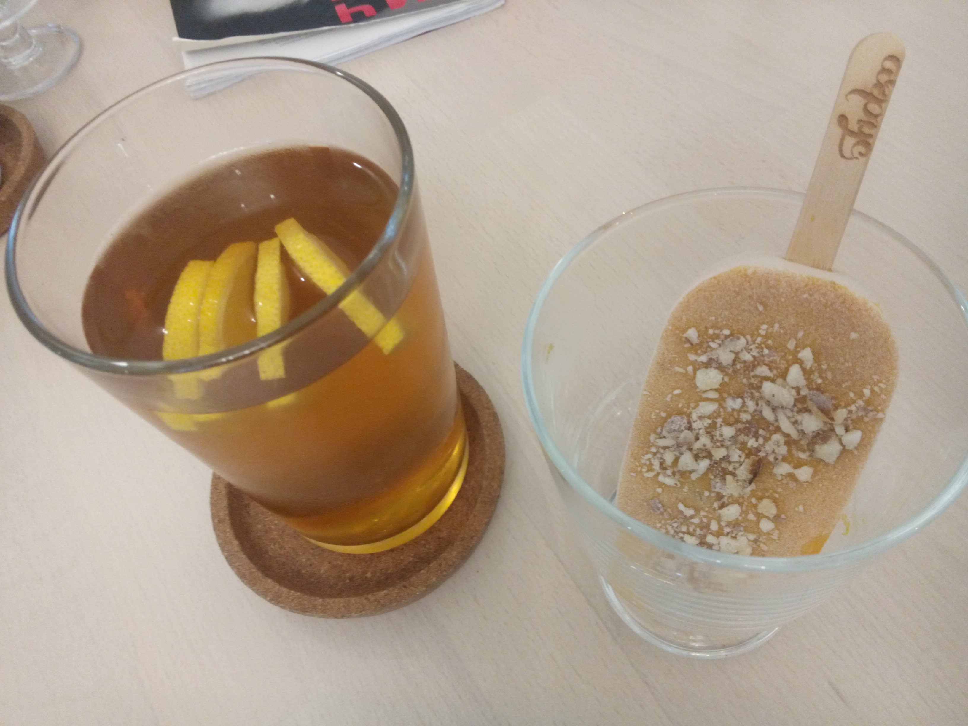A tall glass containing golden tea and lemon slices next to an apricot popsicle covered in chopped nuts