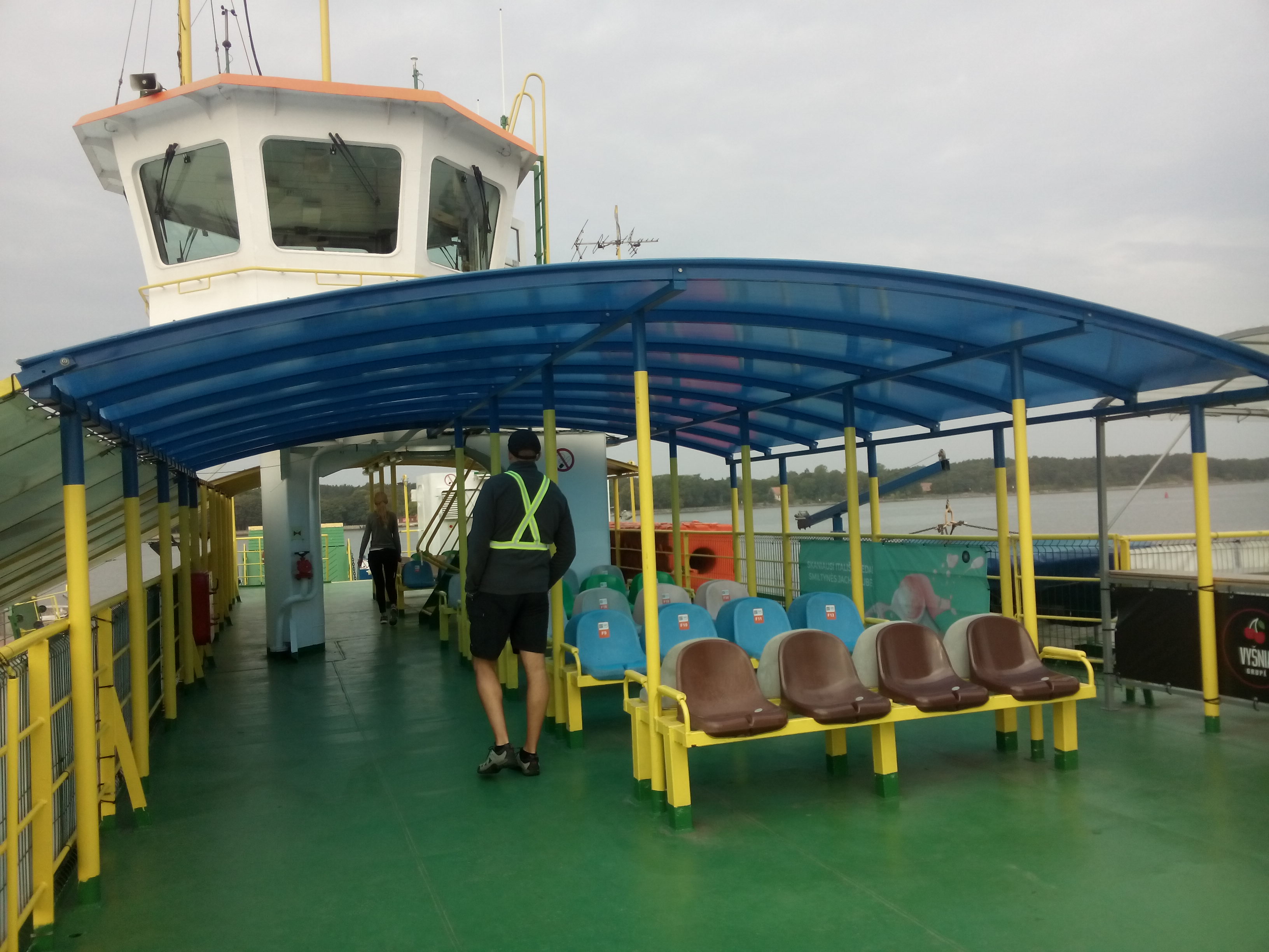A ferry deck with a green floor, plastic blue roof and plastic chairs, with open sides; the cabin is visible at the top and the sky is overcast