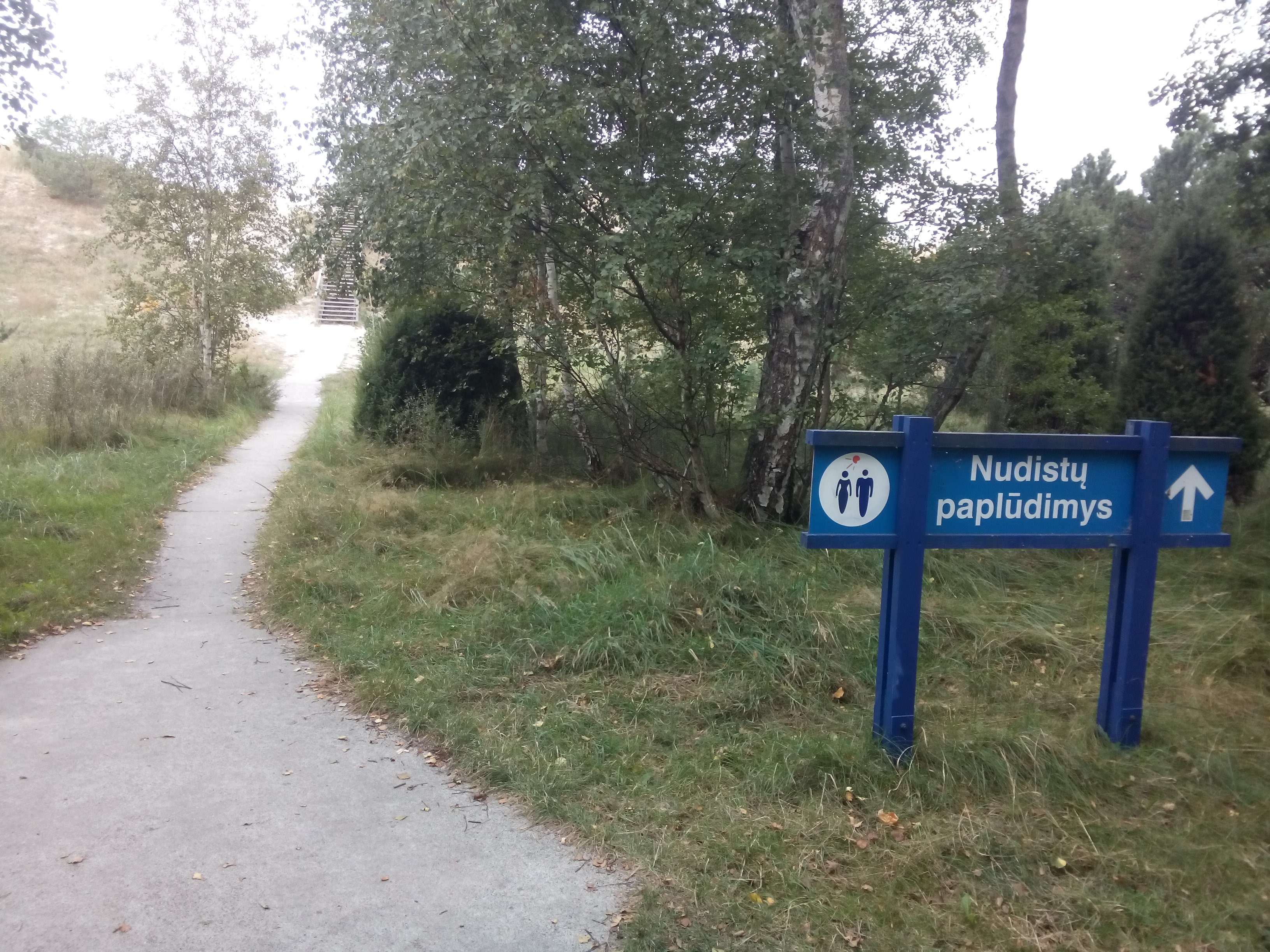 A sandy path leads through the woods; to the right a sign with an arrow says 'nudist beach' in Lithuanian