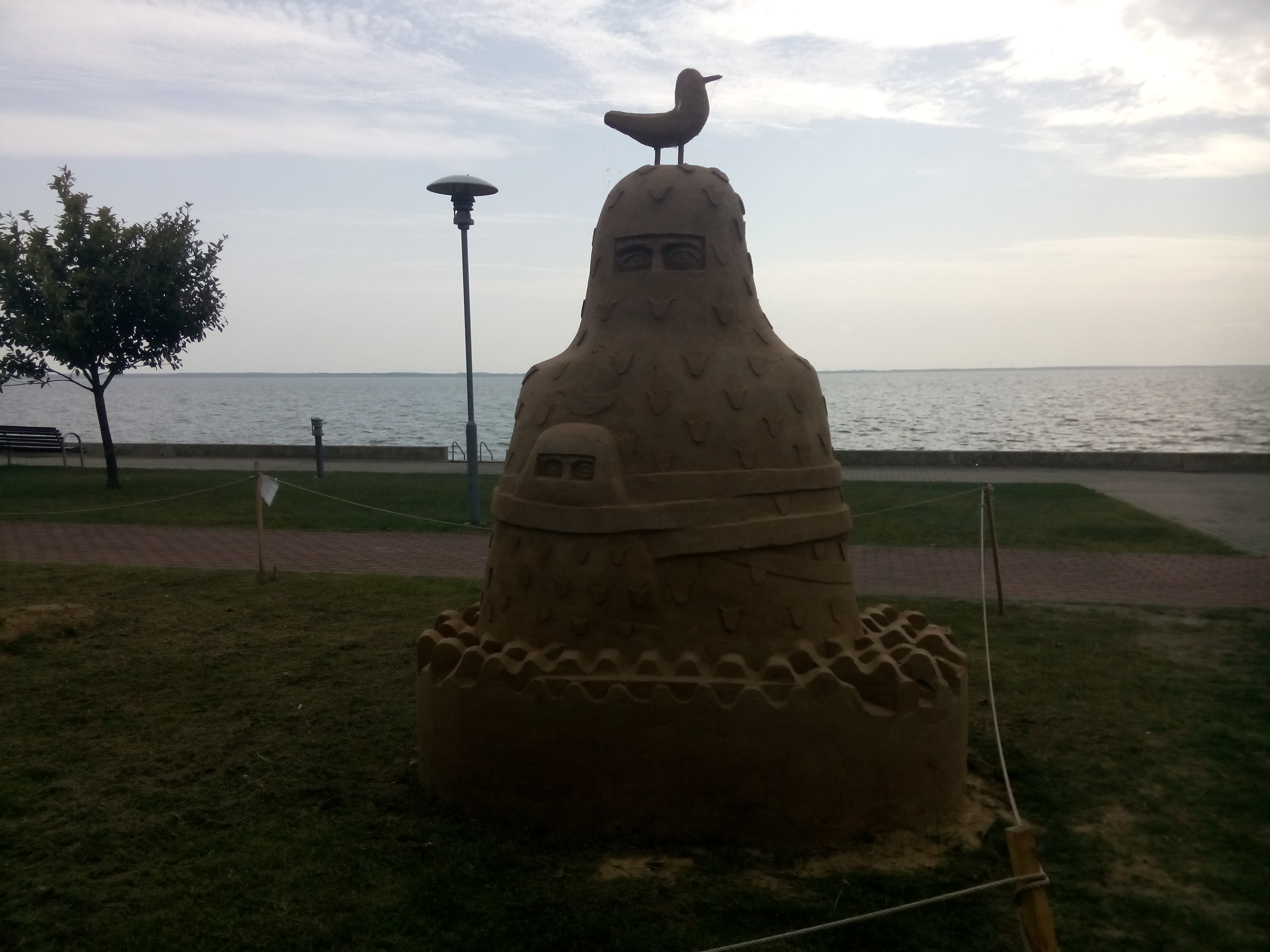 A sand sculpture of two people in veils with a seagull perched on top; the sea in the background