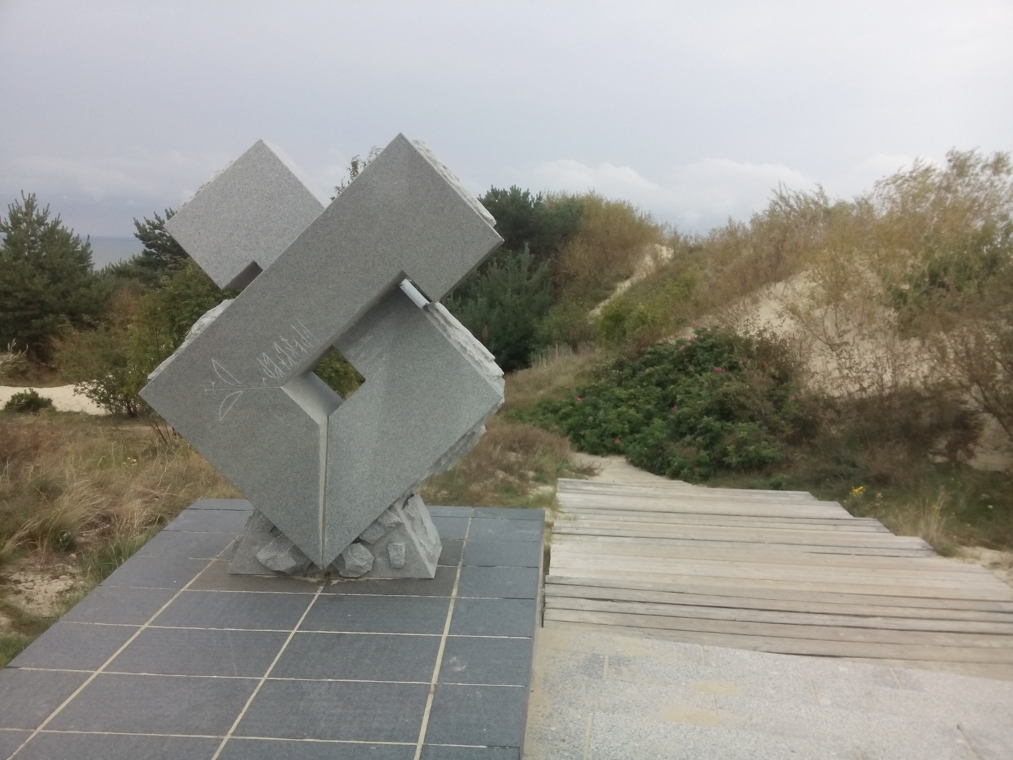 A grey stone sculpture in an abstract diamond shape sits amongst grassy sand dunes