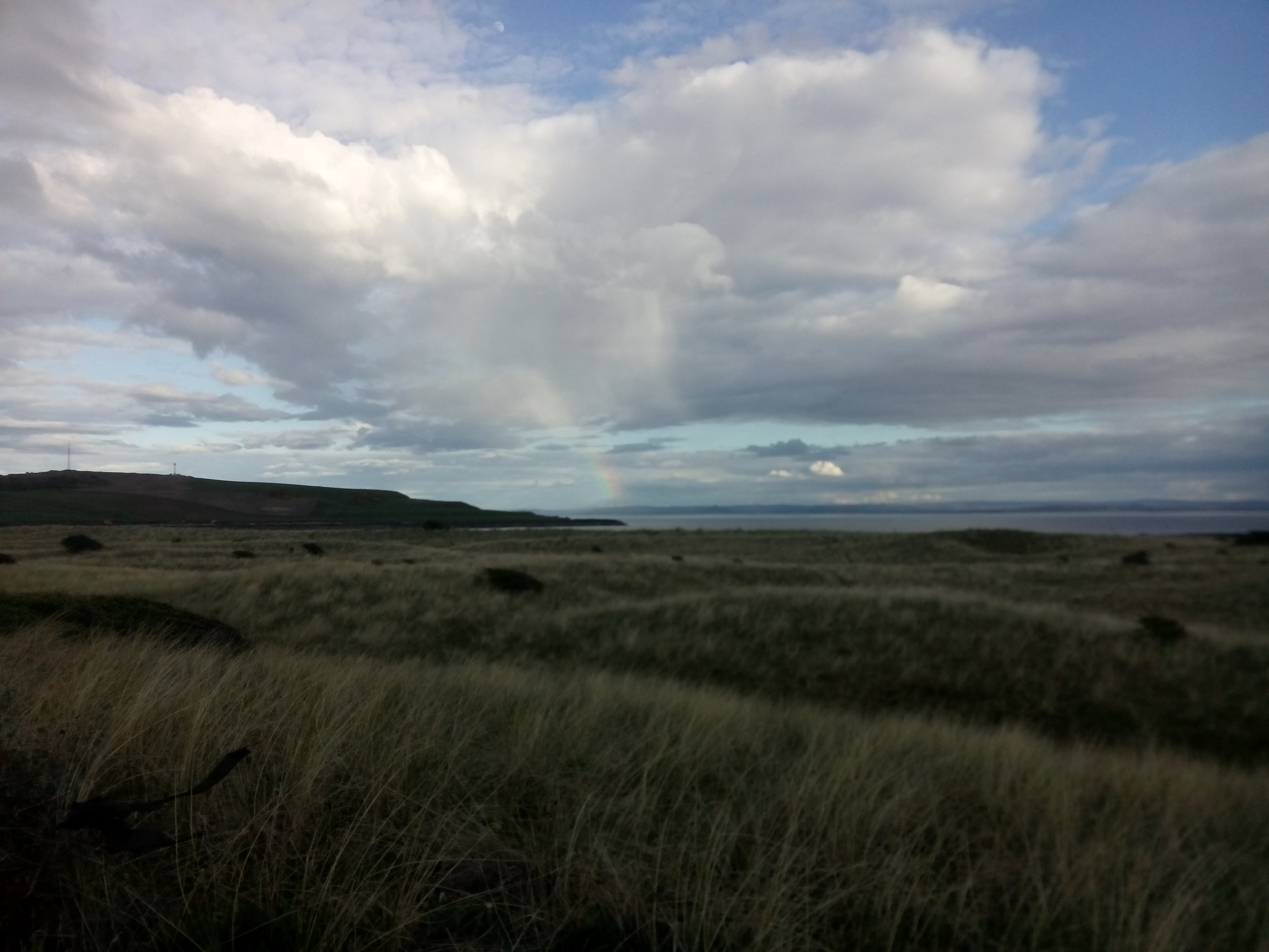 A view across grassy dunes to the sea, where a small rainbow pours down from fluffy clouds