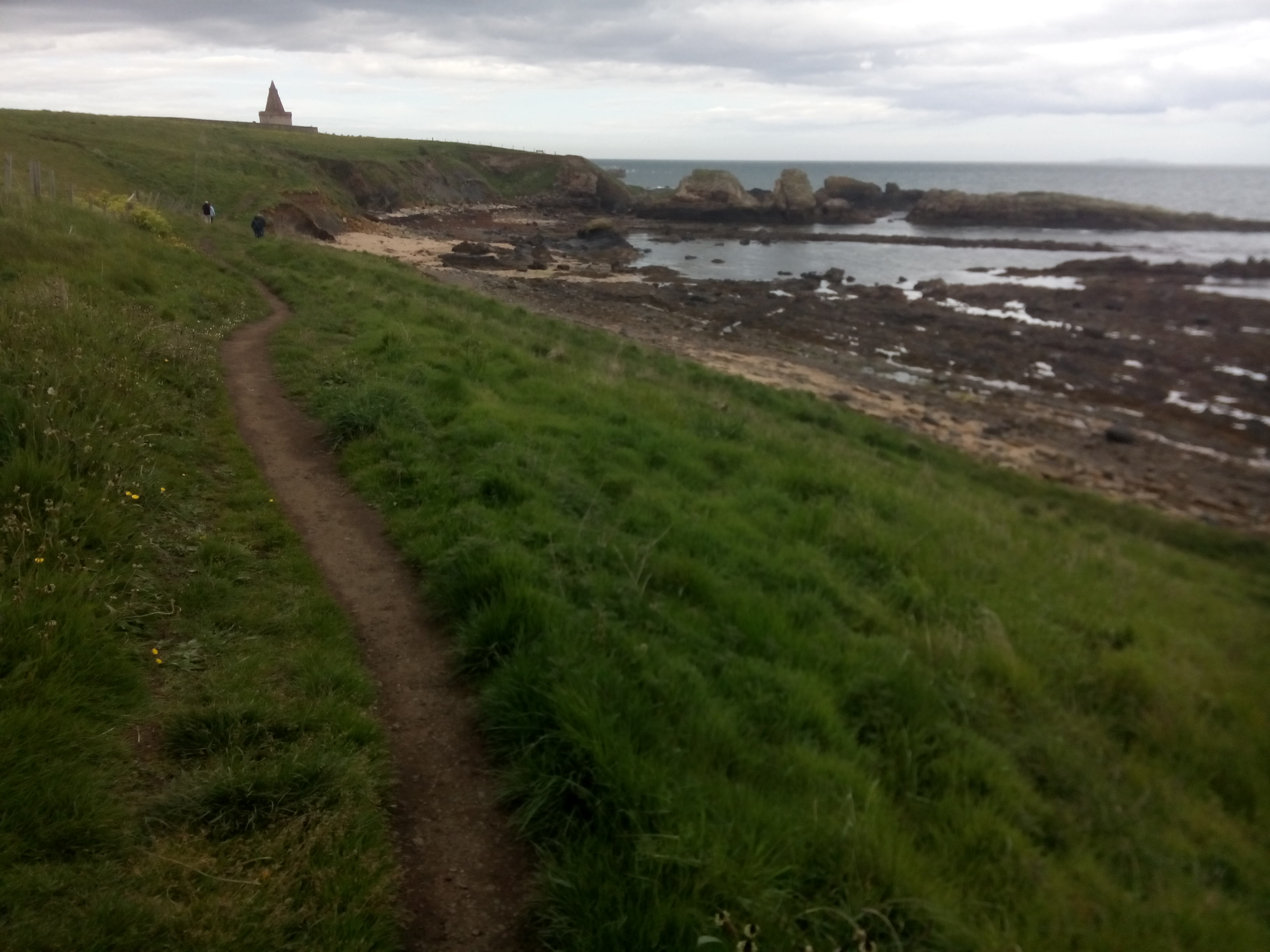 A narrow mud track runs along a grassy verge above a rocky shore