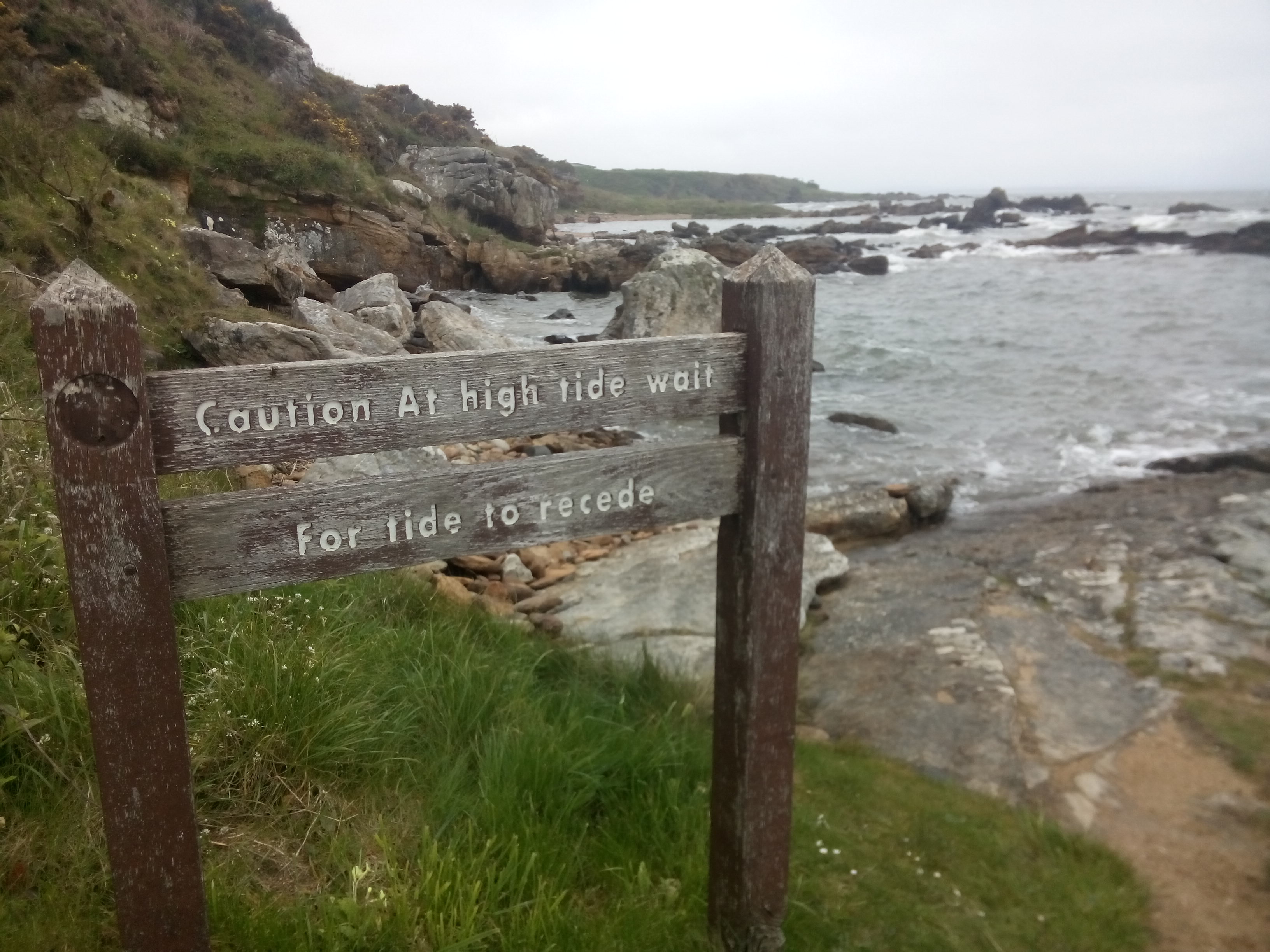 A wooden sign warning about high tides, in front of a rocky cliff that goes directly to the sea