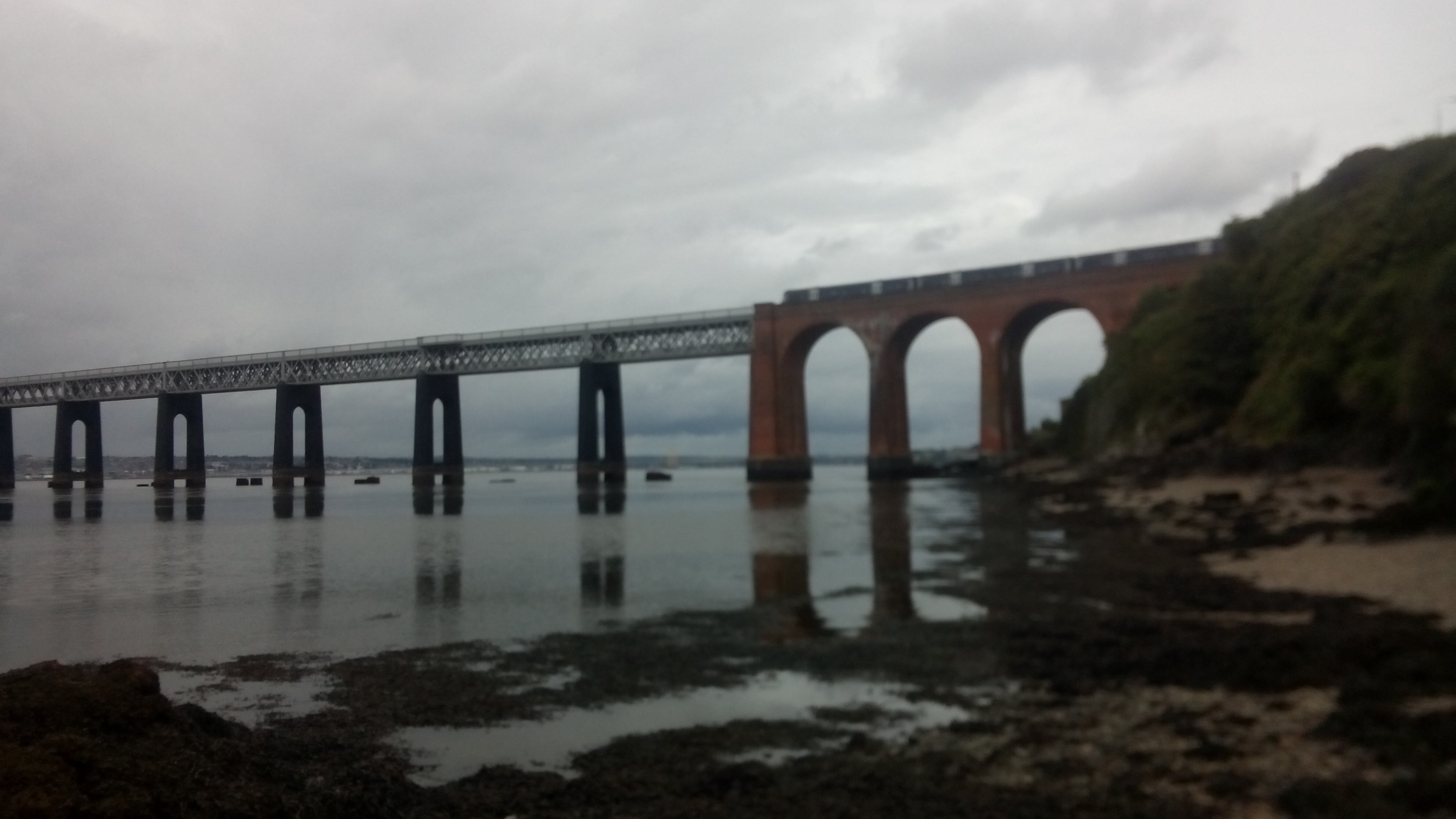 A brick and metal arched railway bridge passes over a rocky beach and water