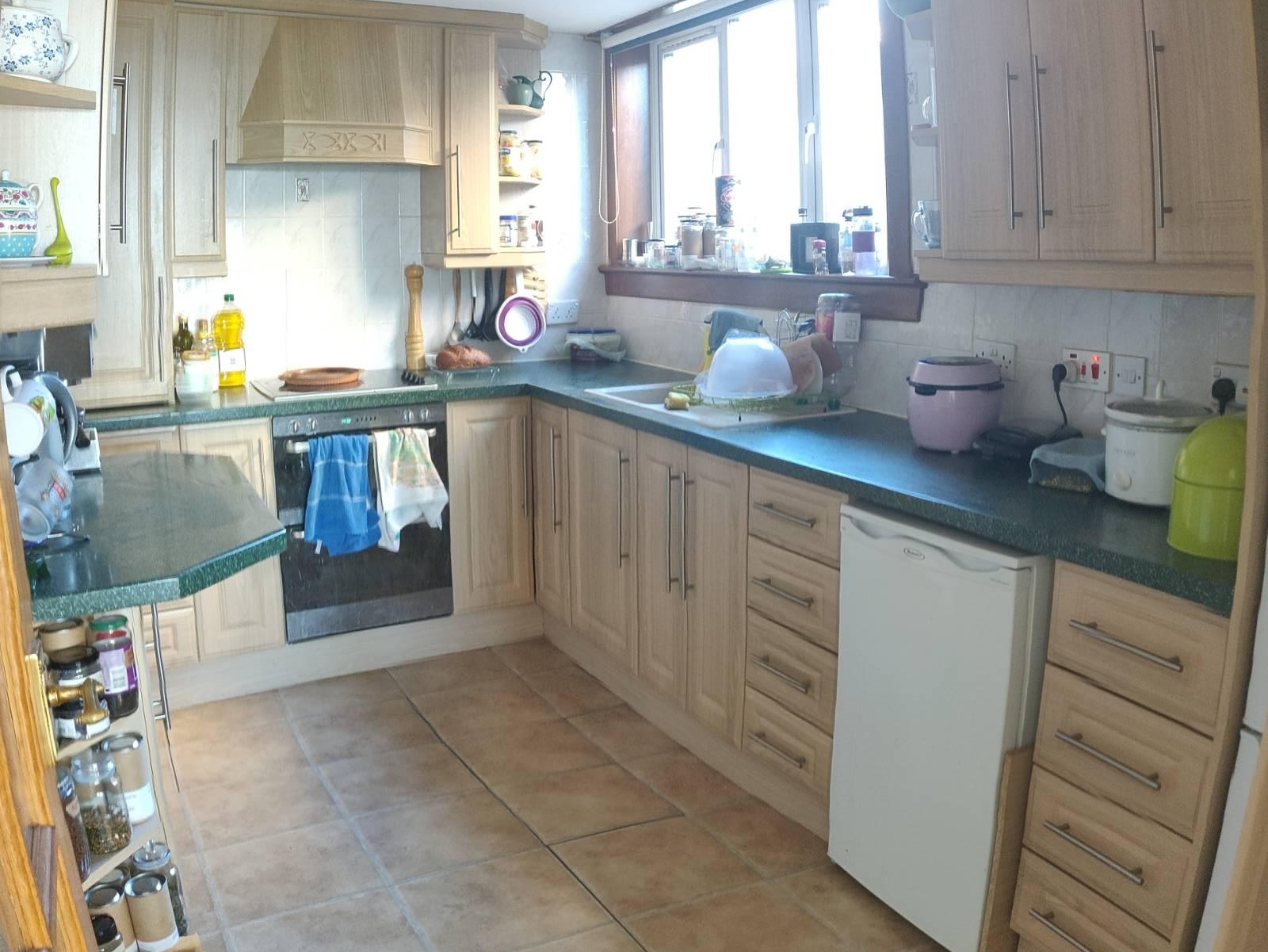 After: A kitchen with green worktops, wood-effect cupboards, shelves full of tea and spaces, utensils hanging